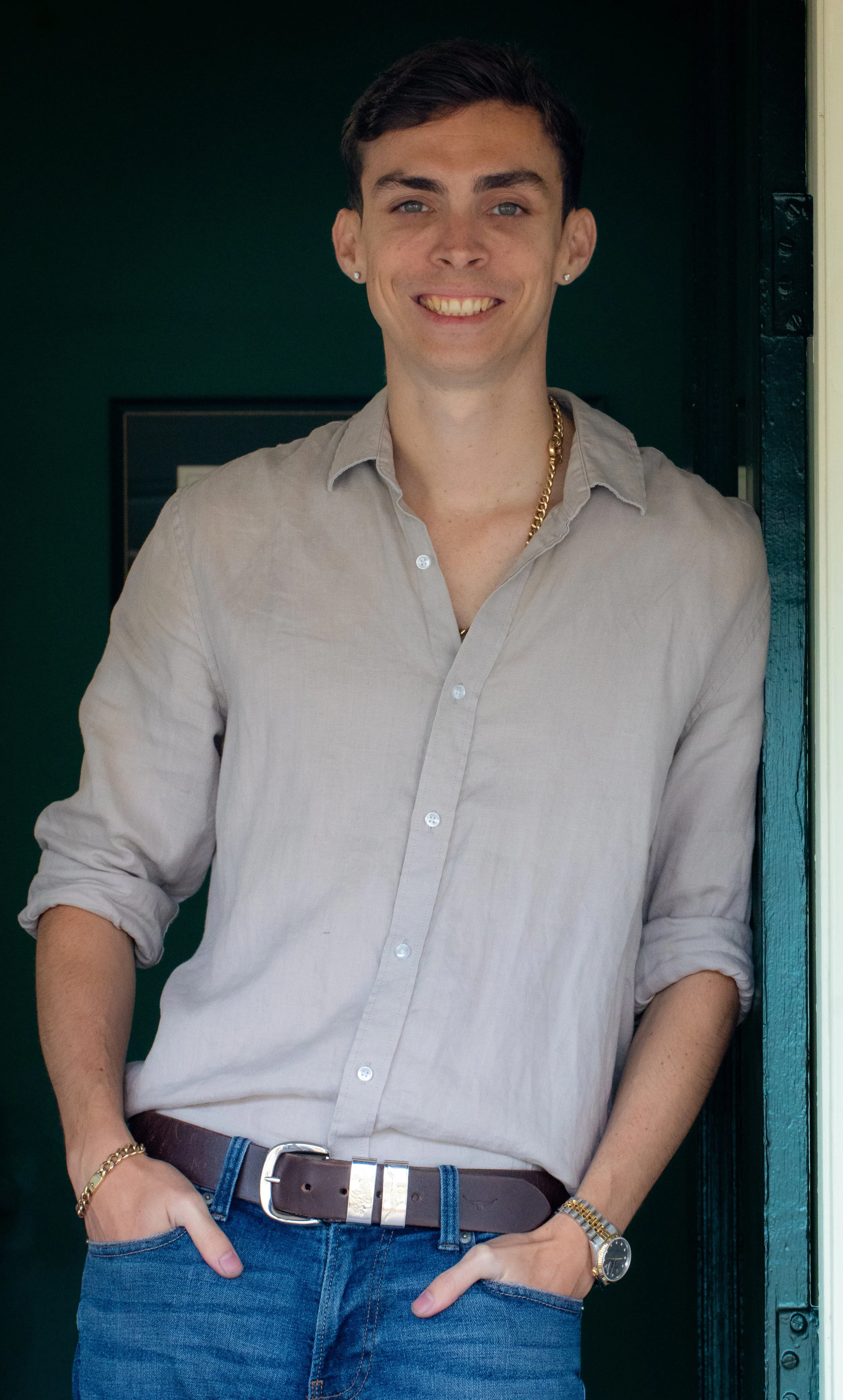 Young man with dark hair, wearing a light beige button-up shirt with rolled-up sleeves, blue jeans, gold chain necklace, leather belt, and a watch. He is smiling and standing with his hands in his pockets in front of a dark green door.