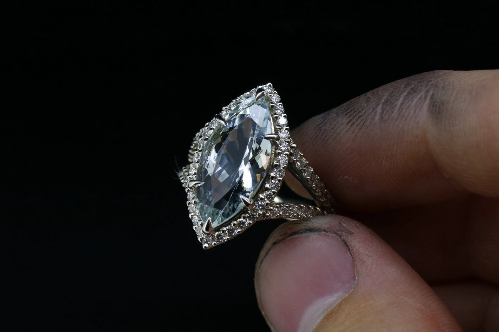 Close-up of a large, oval clear gemstone ring with smaller stones around it, held between fingers against a black background.