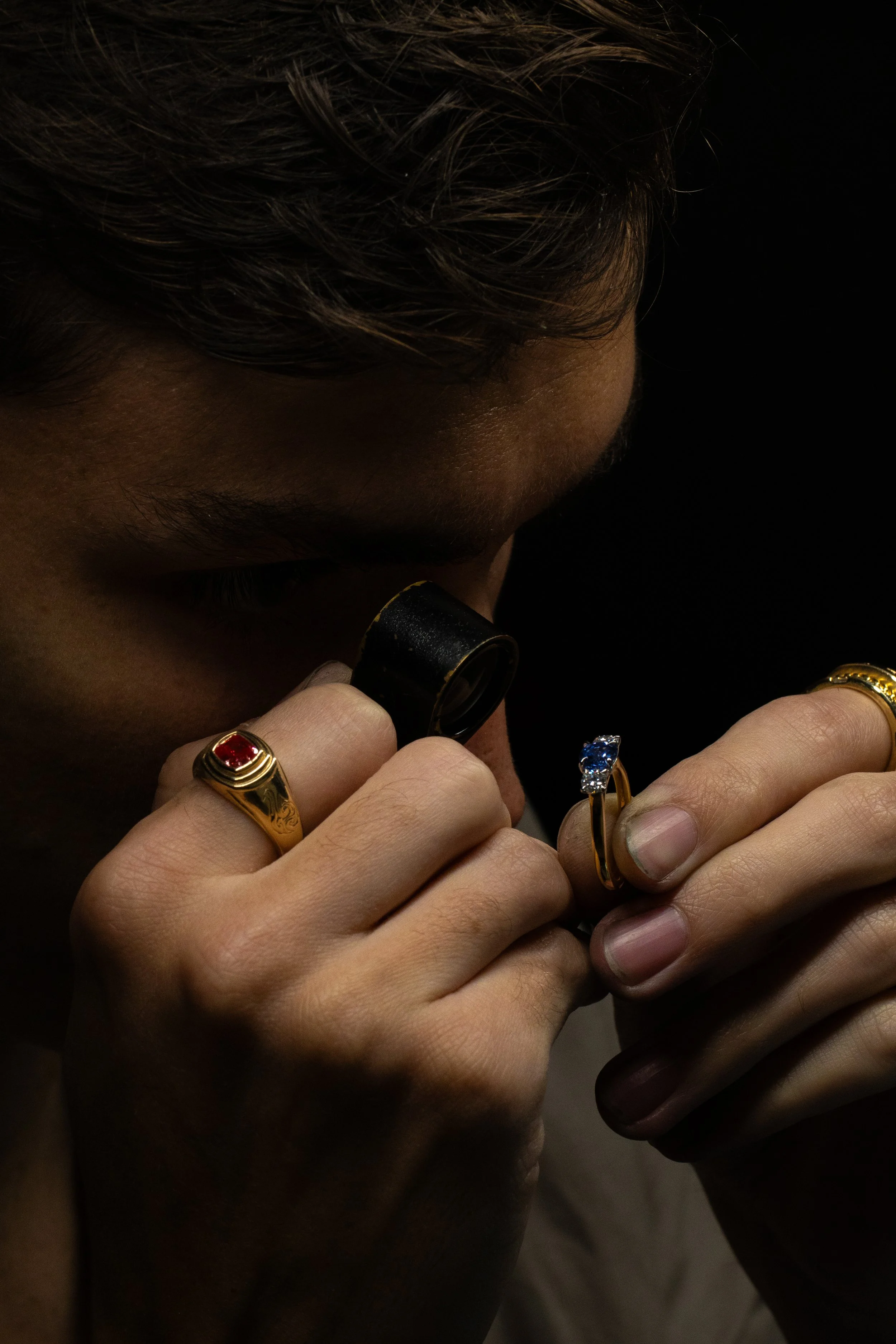 Person examining jewelry with a magnifying glass, wearing multiple rings, including a red gemstone and a blue gemstone ring.