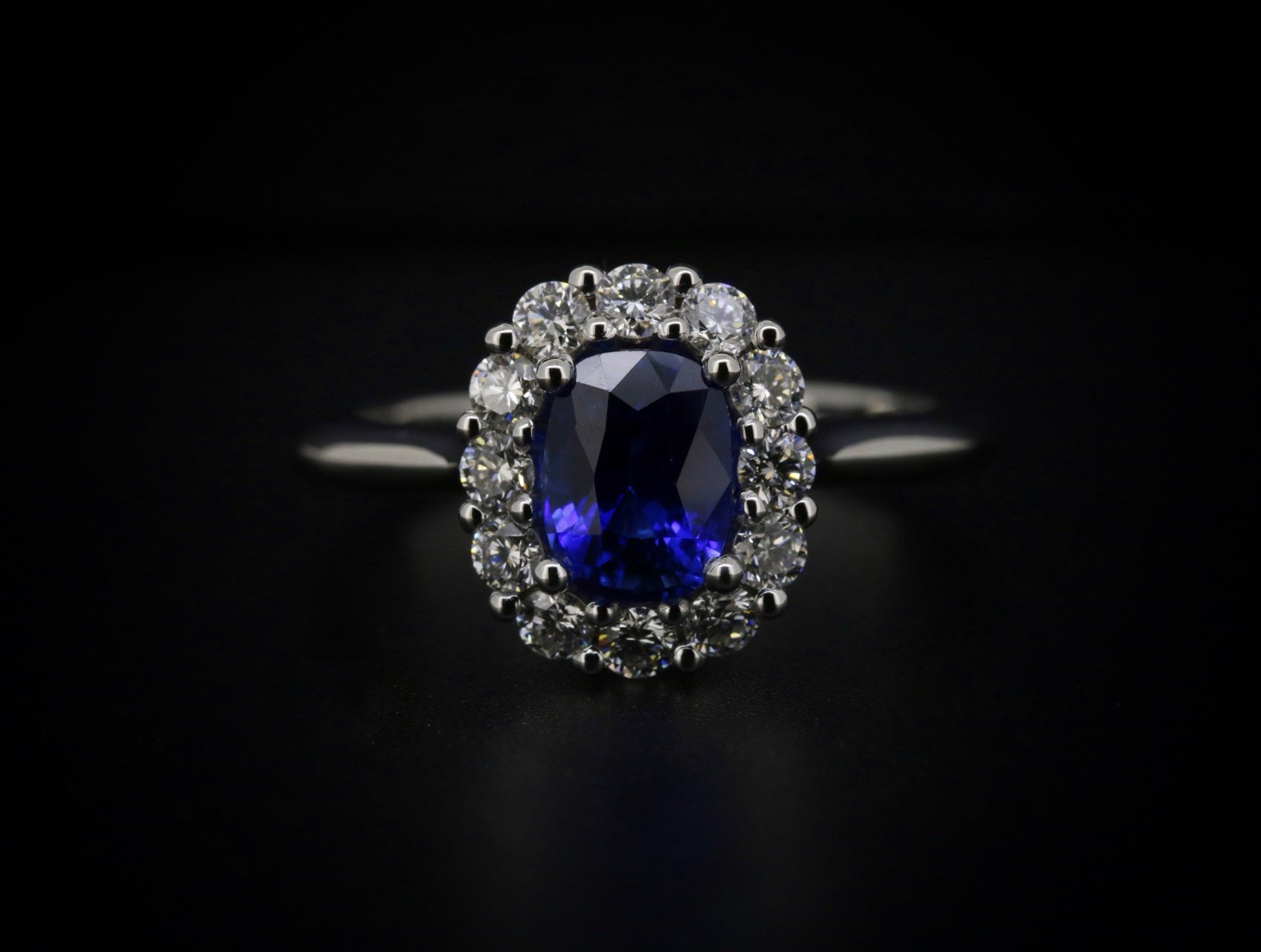 A silver ring featuring a large, oval-shaped, deep blue gemstone surrounded by a halo of smaller clear diamonds, against a black background.
