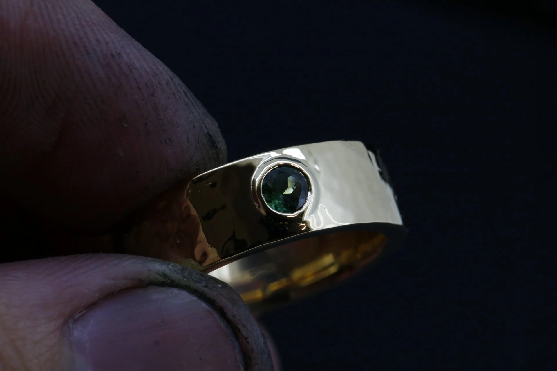 Close-up of a hand holding a shiny silver ring with a dark green gemstone against a black background.