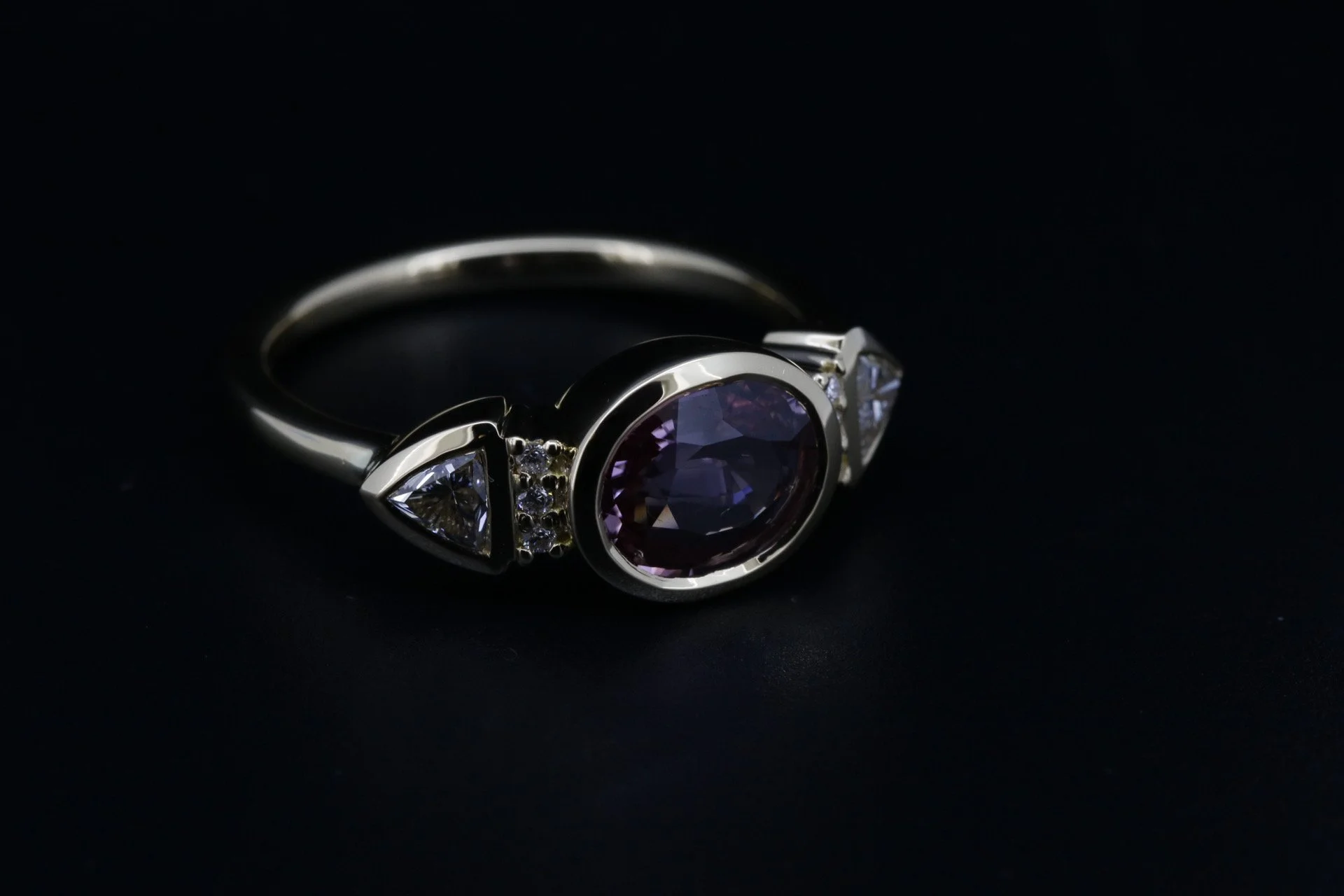 A silver ring with a large central dark purple gemstone and two smaller clear gemstones on each side, set against a black background.