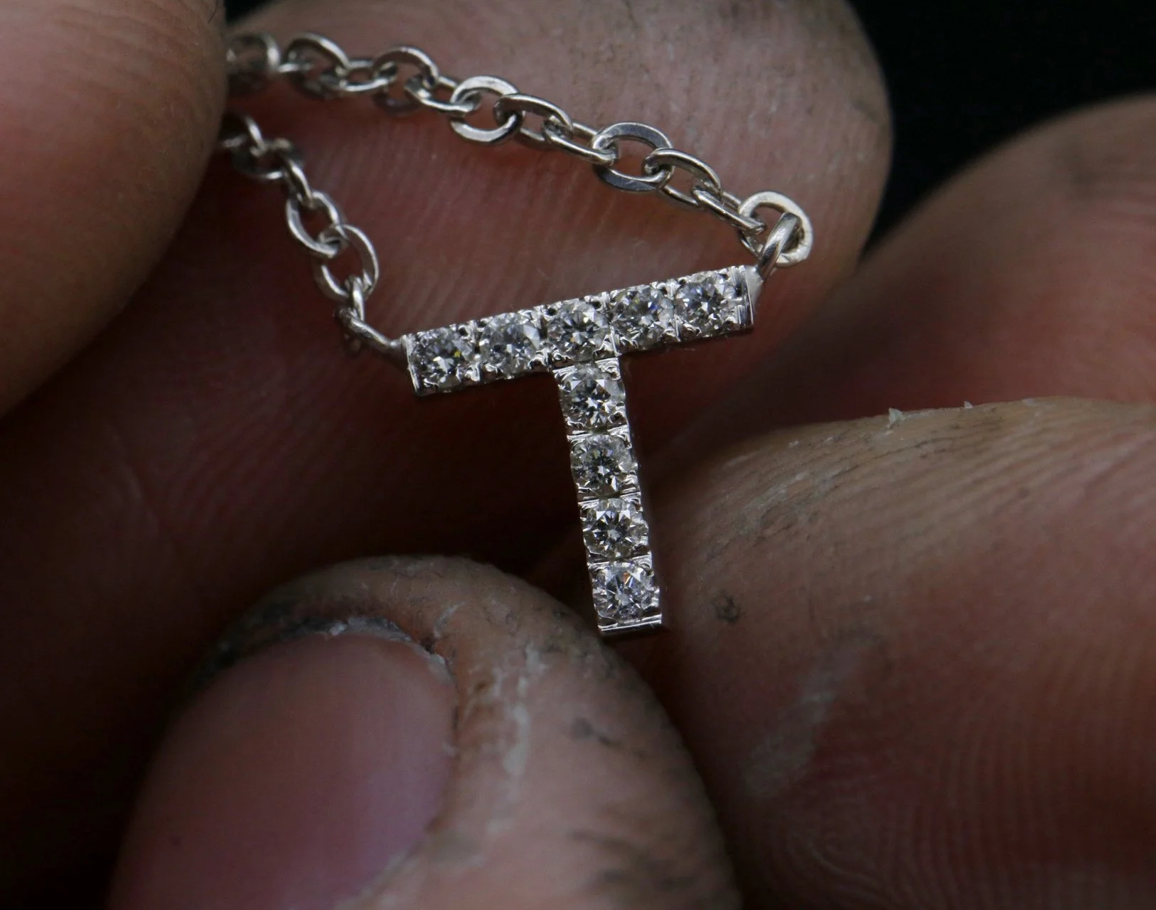 A close-up of a person's fingers holding a silver chain necklace with a letter 'T' pendant adorned with small diamonds.