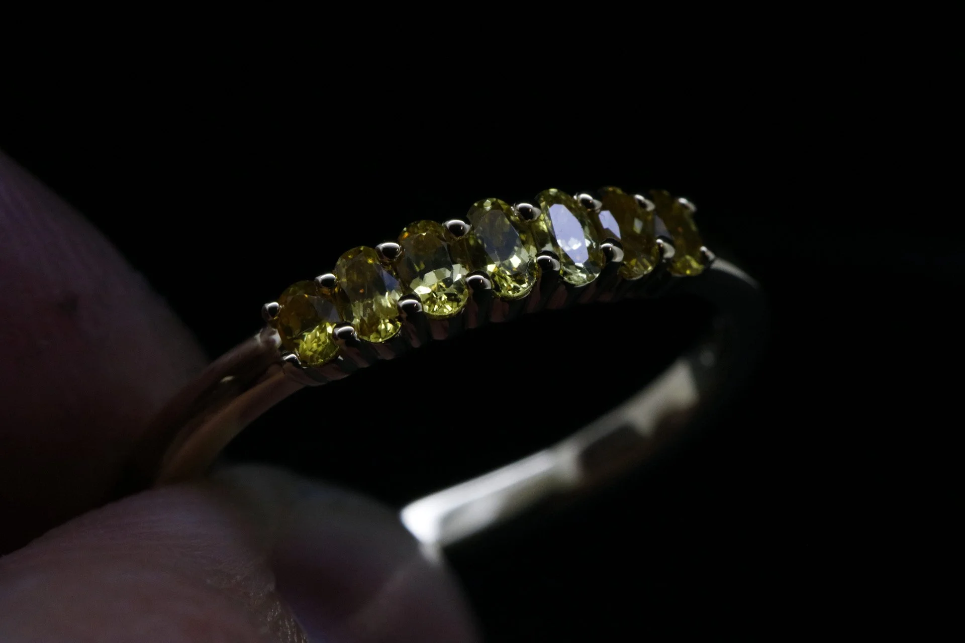 A ring with a row of yellow gemstones set in a silver band on a person's finger, with a dark background.