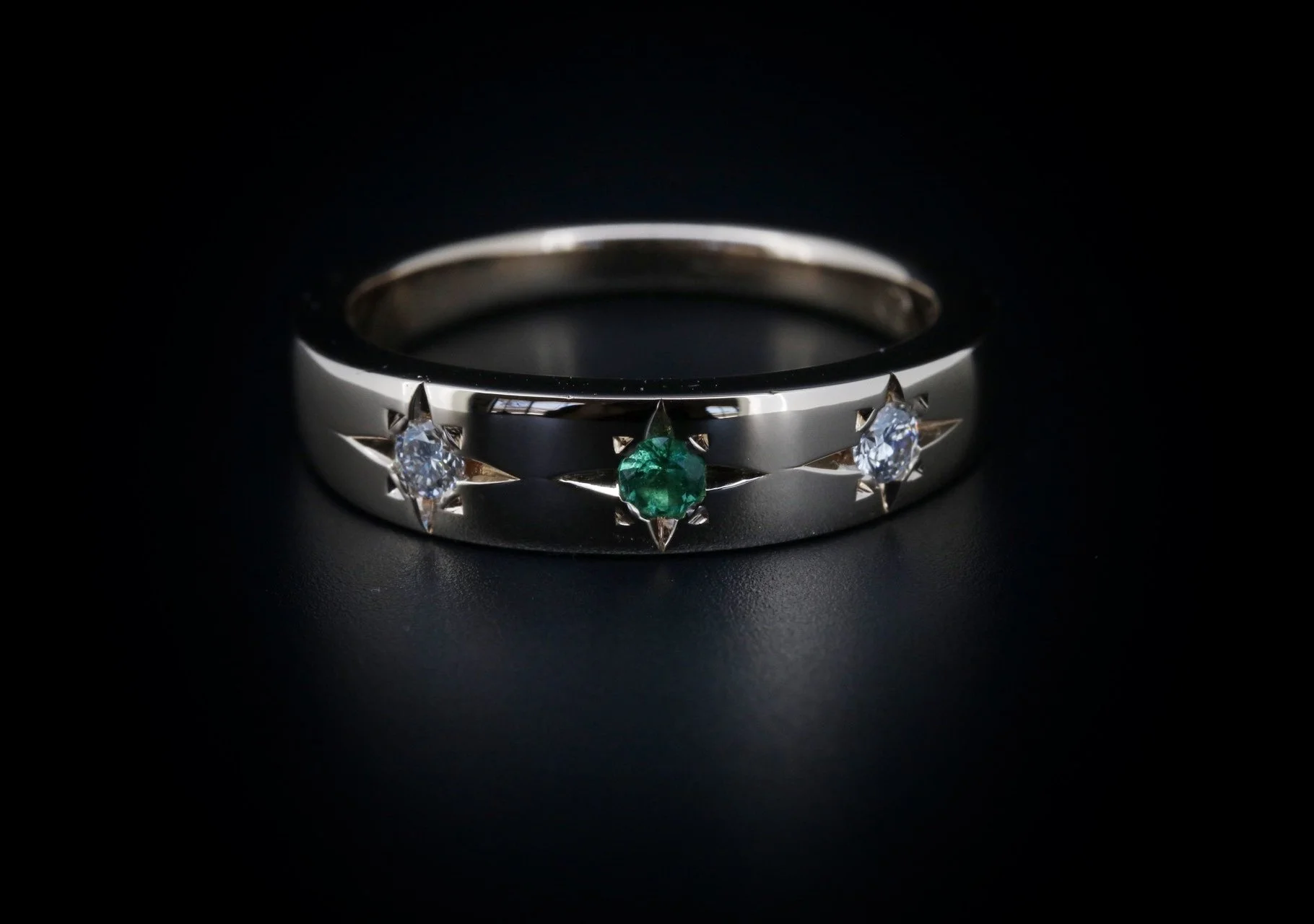 A silver ring with three prong-set gemstones, including one central green gemstone and two clear gemstones, against a black background.