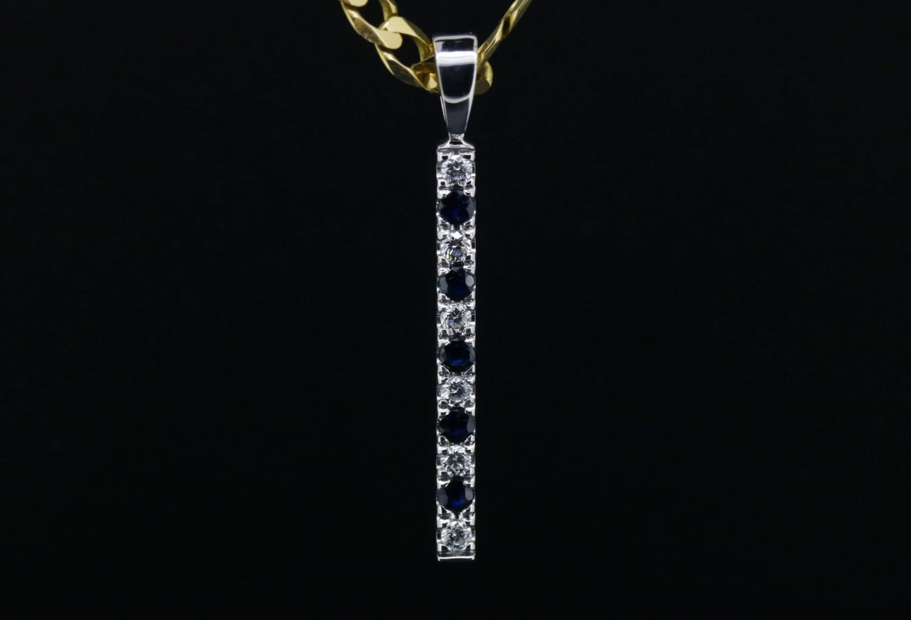 A vertical pendant necklace featuring a row of alternating diamonds and dark blue sapphires, hanging from a gold chain against a black background.