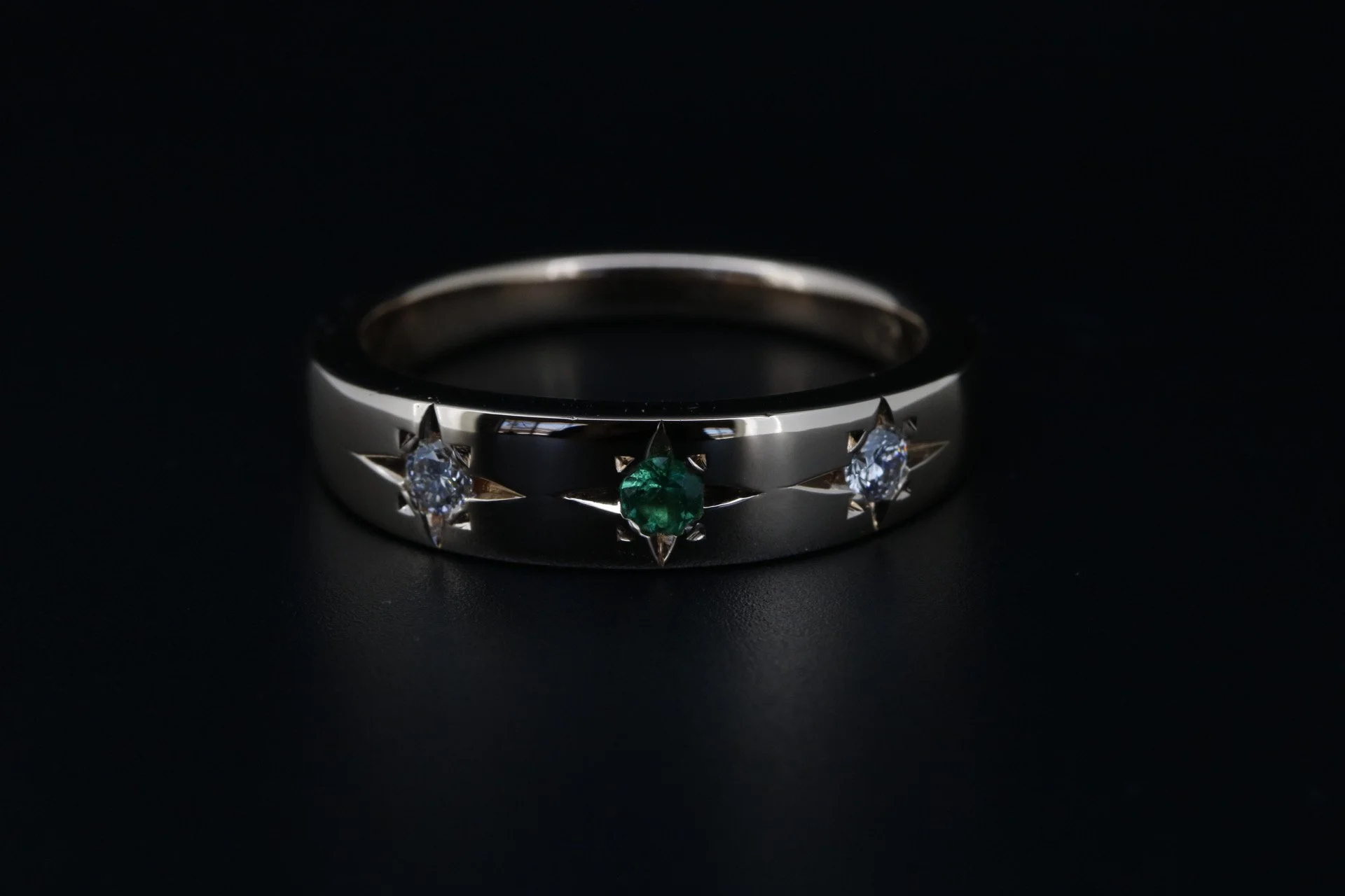 A silver ring with three gemstones: two clear diamonds and one green gemstone, set against a dark background.