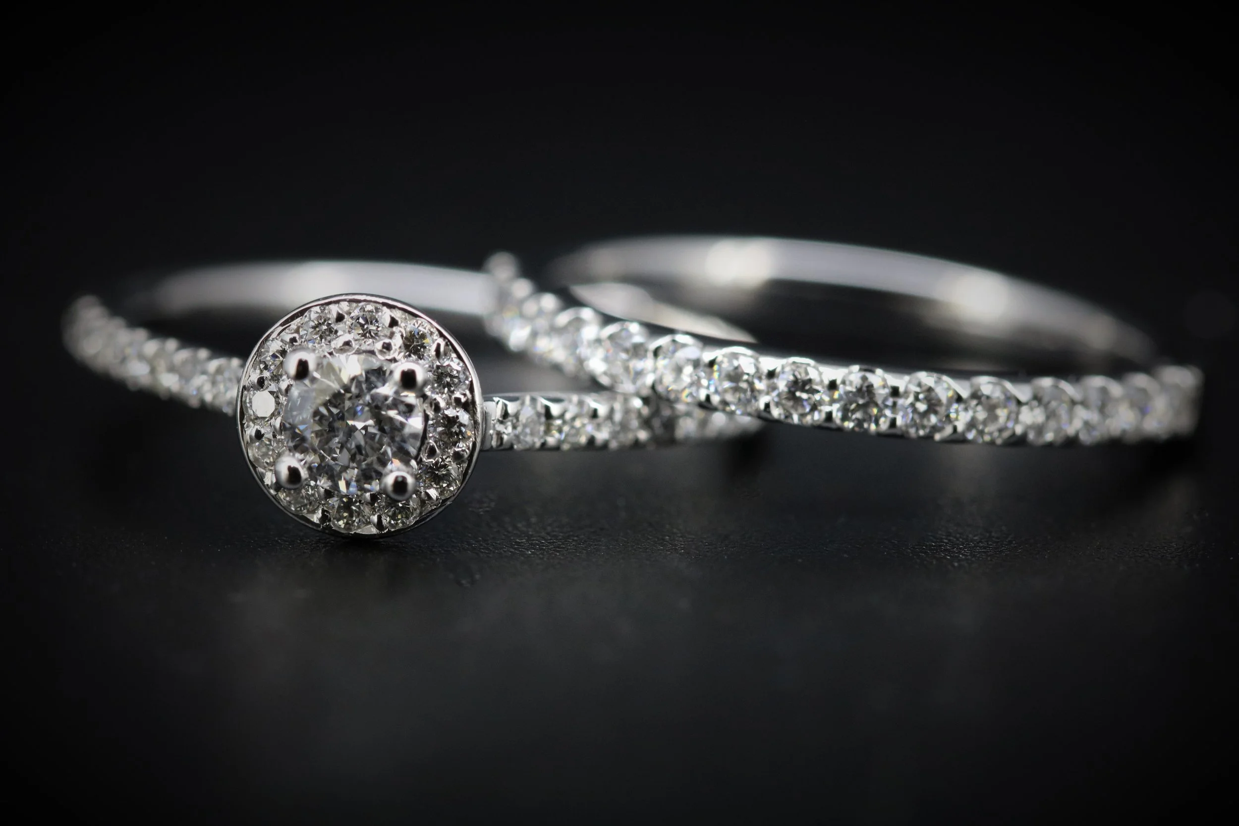 Close-up of two silver rings with diamonds, one with a larger round diamond in the center surrounded by smaller diamonds, on a black background.