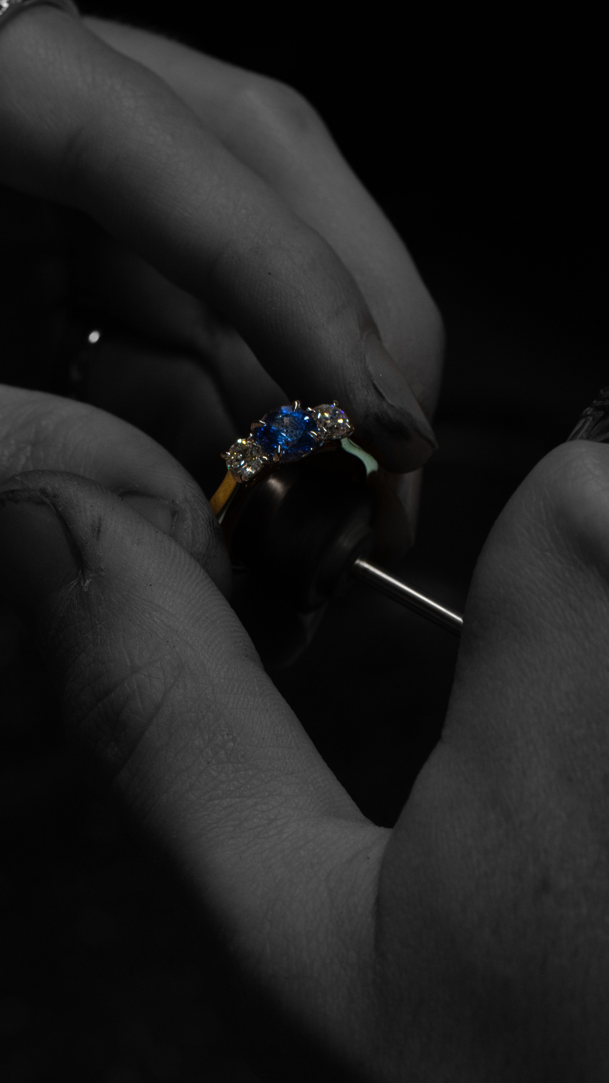 A jewelry maker's hands, covered in protective gloves, polishing a gold ring with a blue gemstone and additional smaller stones.