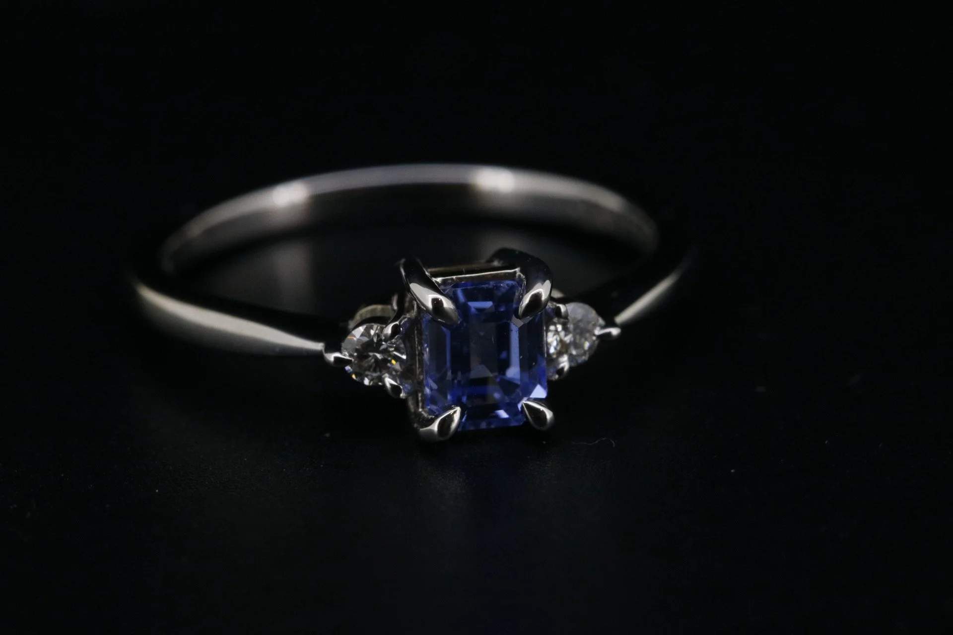 Silver ring with a blue rectangular gemstone flanked by two smaller clear gemstones, set against a black background.