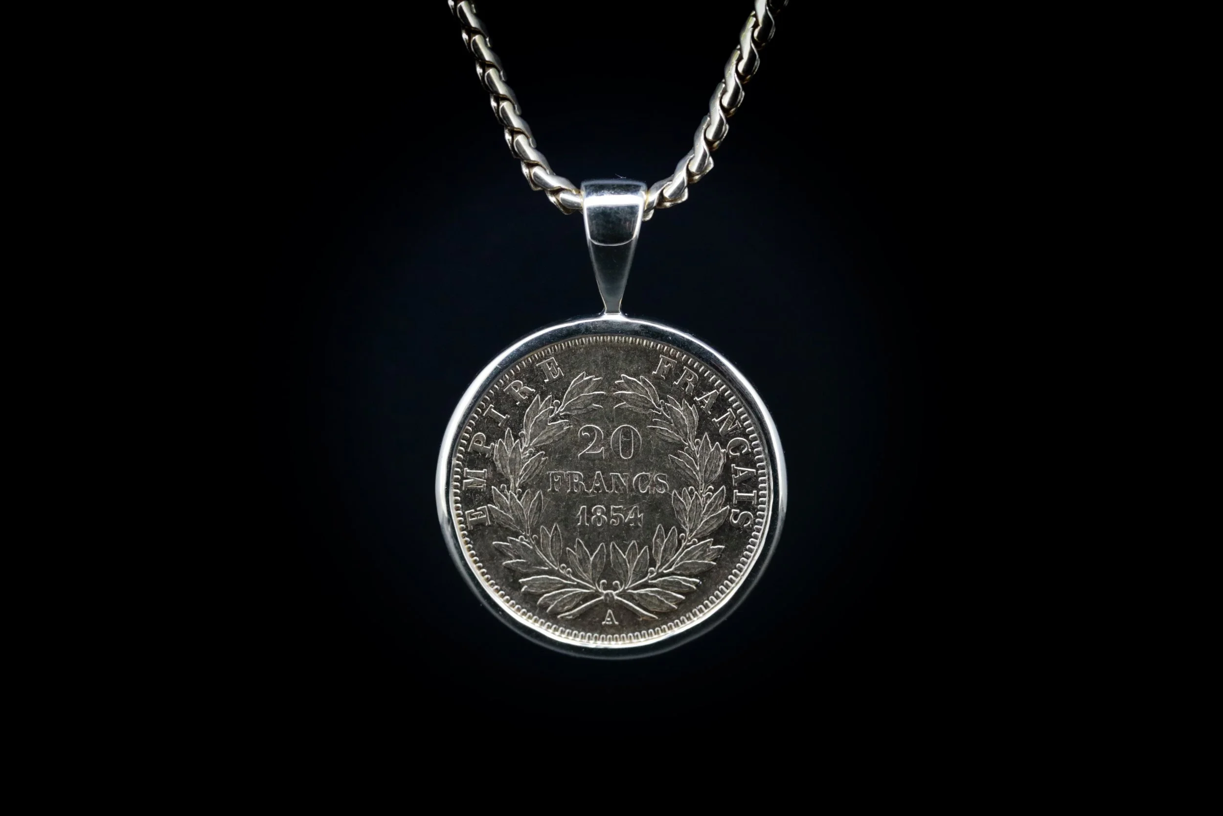 A silver pendant necklace with a coin featuring French text and a decorative laurel wreath design. The chain is thick and twisted, and the background is black.