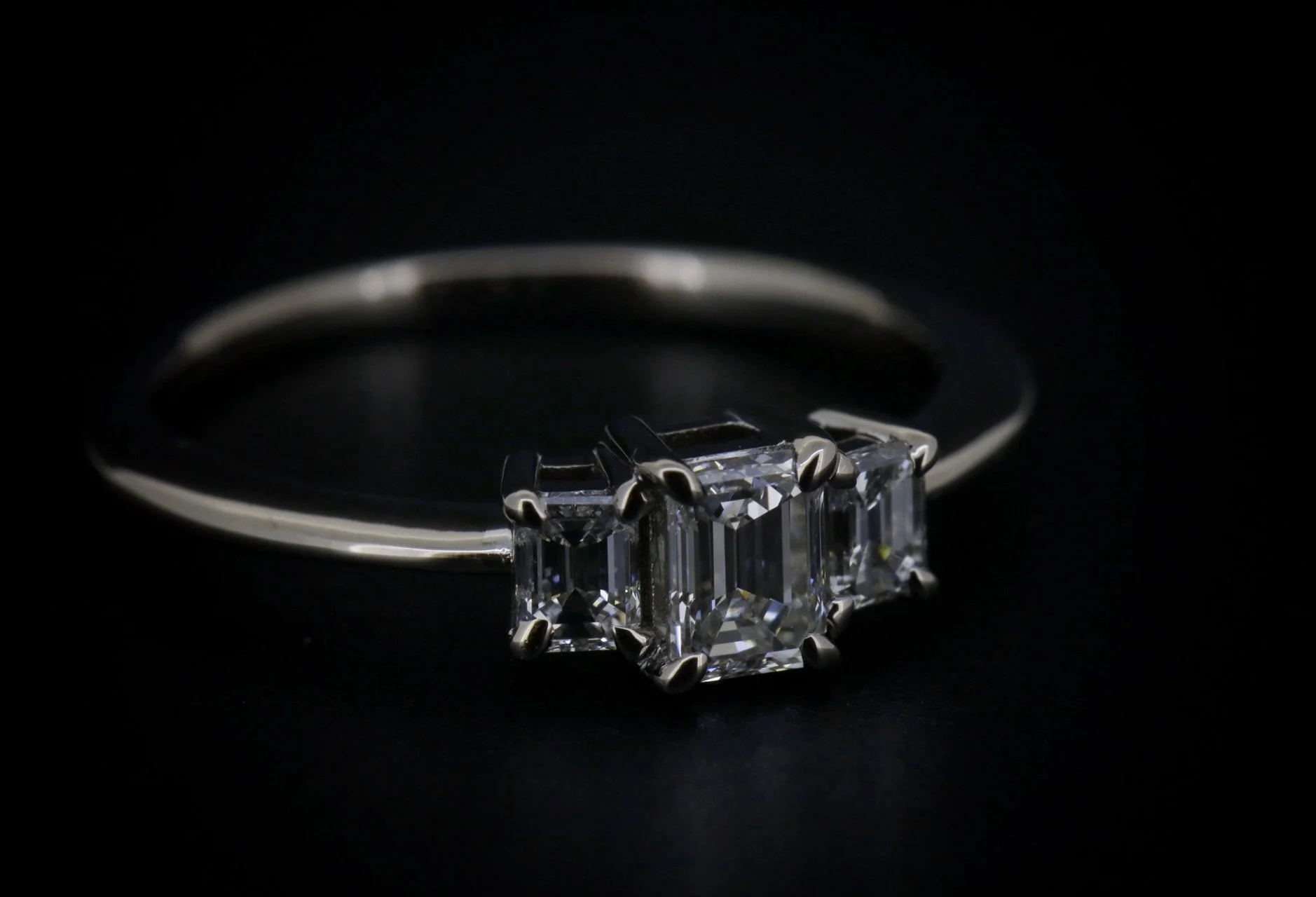 A silver ring with three large emerald-cut diamonds set in a row on a black background.