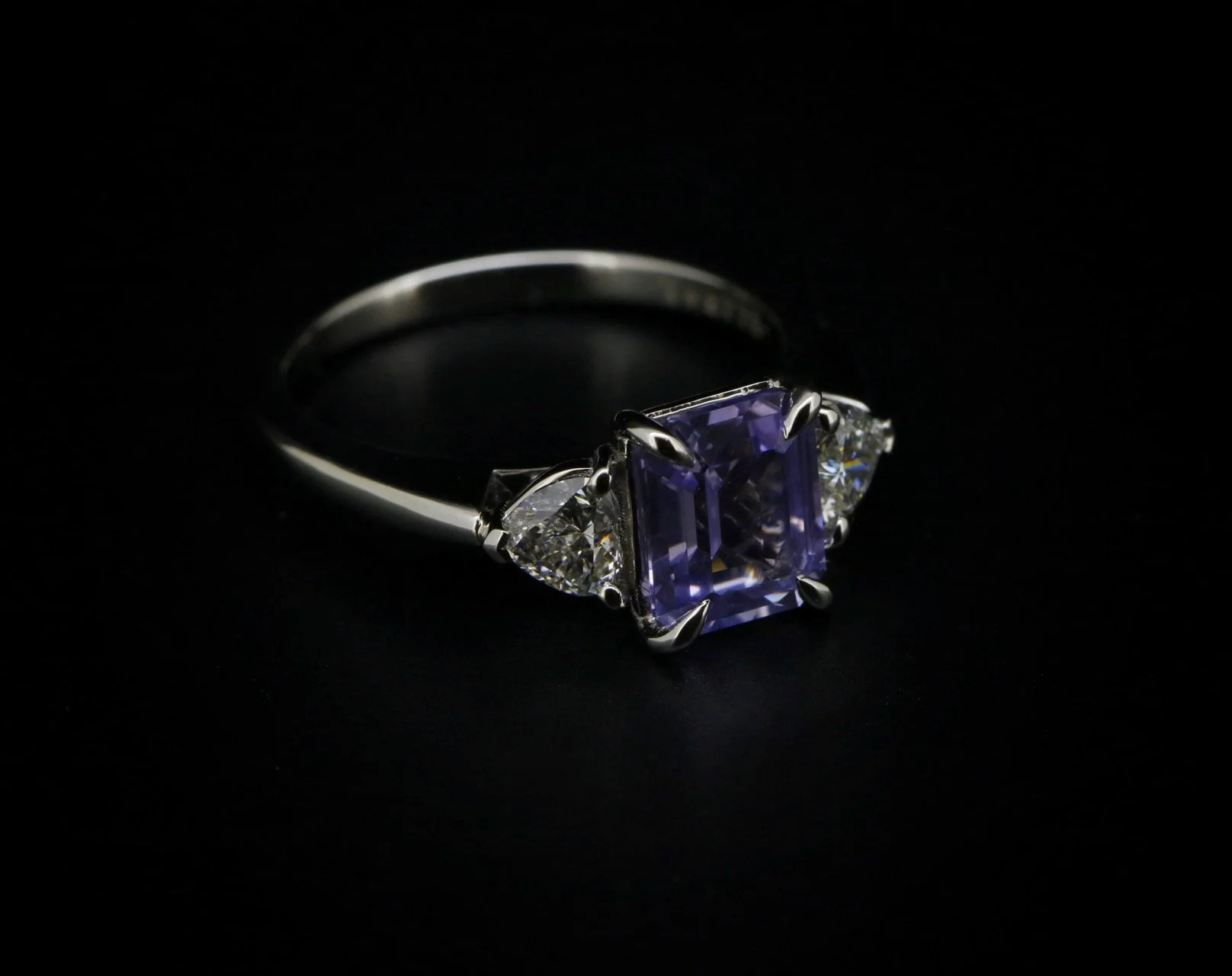 A silver ring with a large purple square gemstone flanked by two clear triangular diamonds on a black background.