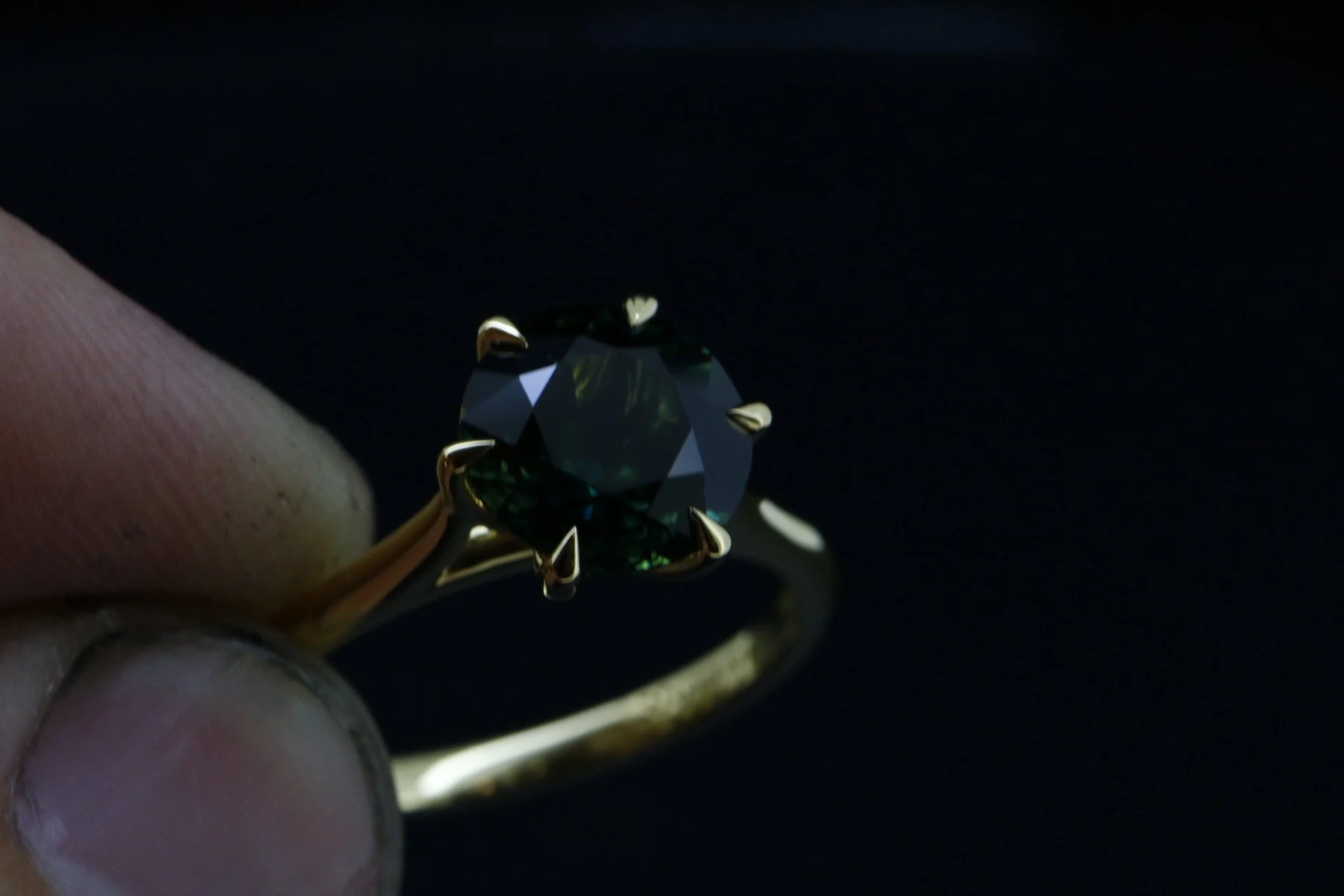 Close-up of a gold ring with a large, dark green gemstone set in a prong setting, held between fingers against a black background.