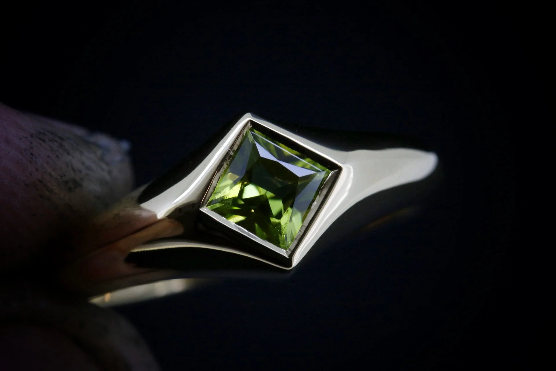 Close-up of a silver ring with a green square-cut gemstone set in a bezel setting against a dark background.