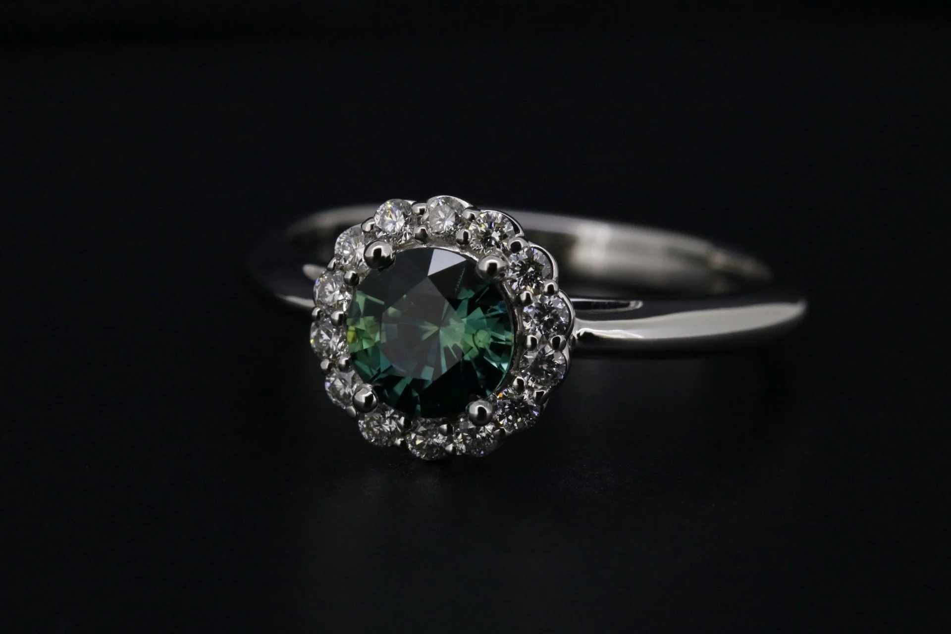 Silver ring with a large greenish-blue central gemstone surrounded by small clear diamonds on a black background.