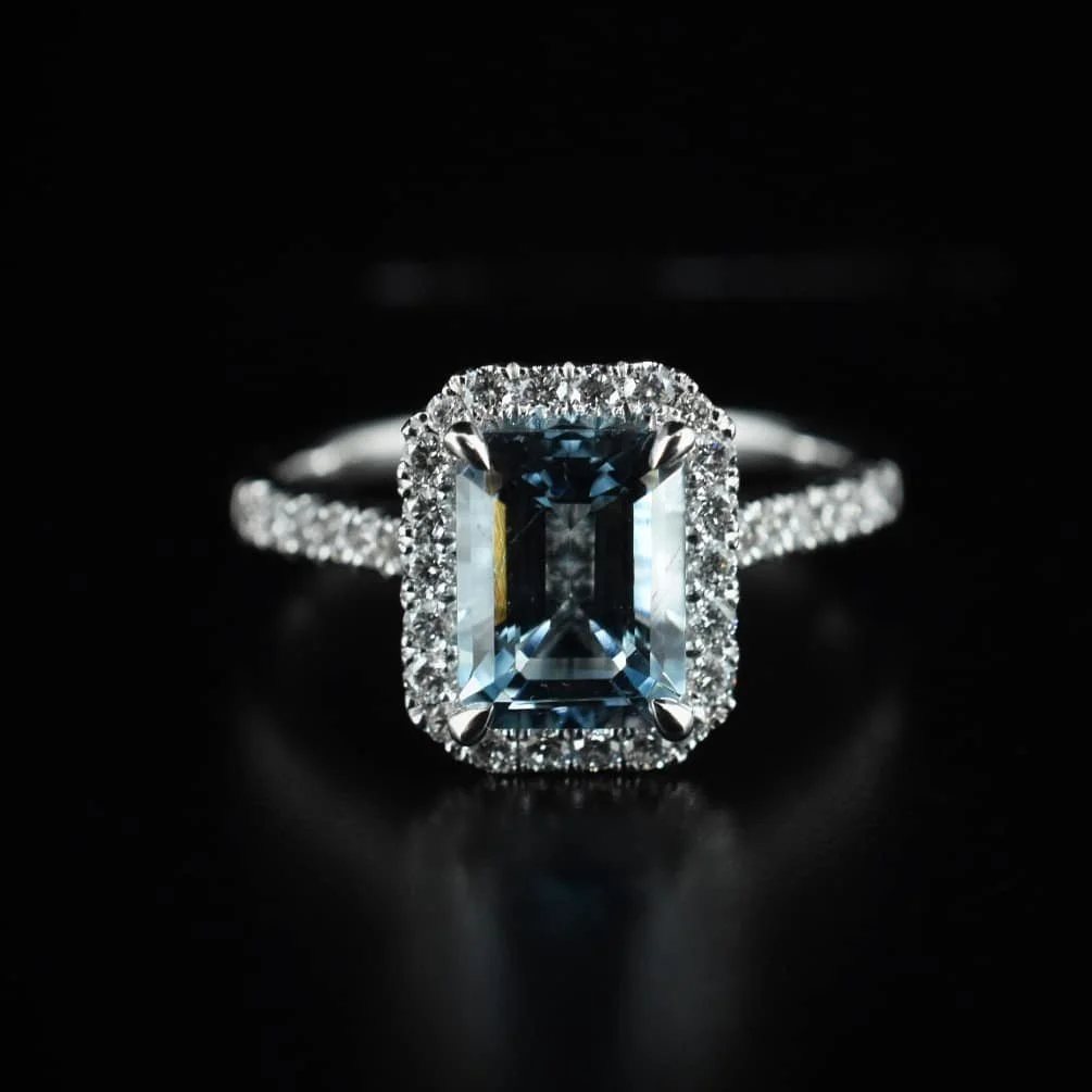 A silver ring with an emerald-cut blue gemstone surrounded by small diamonds on a black background.