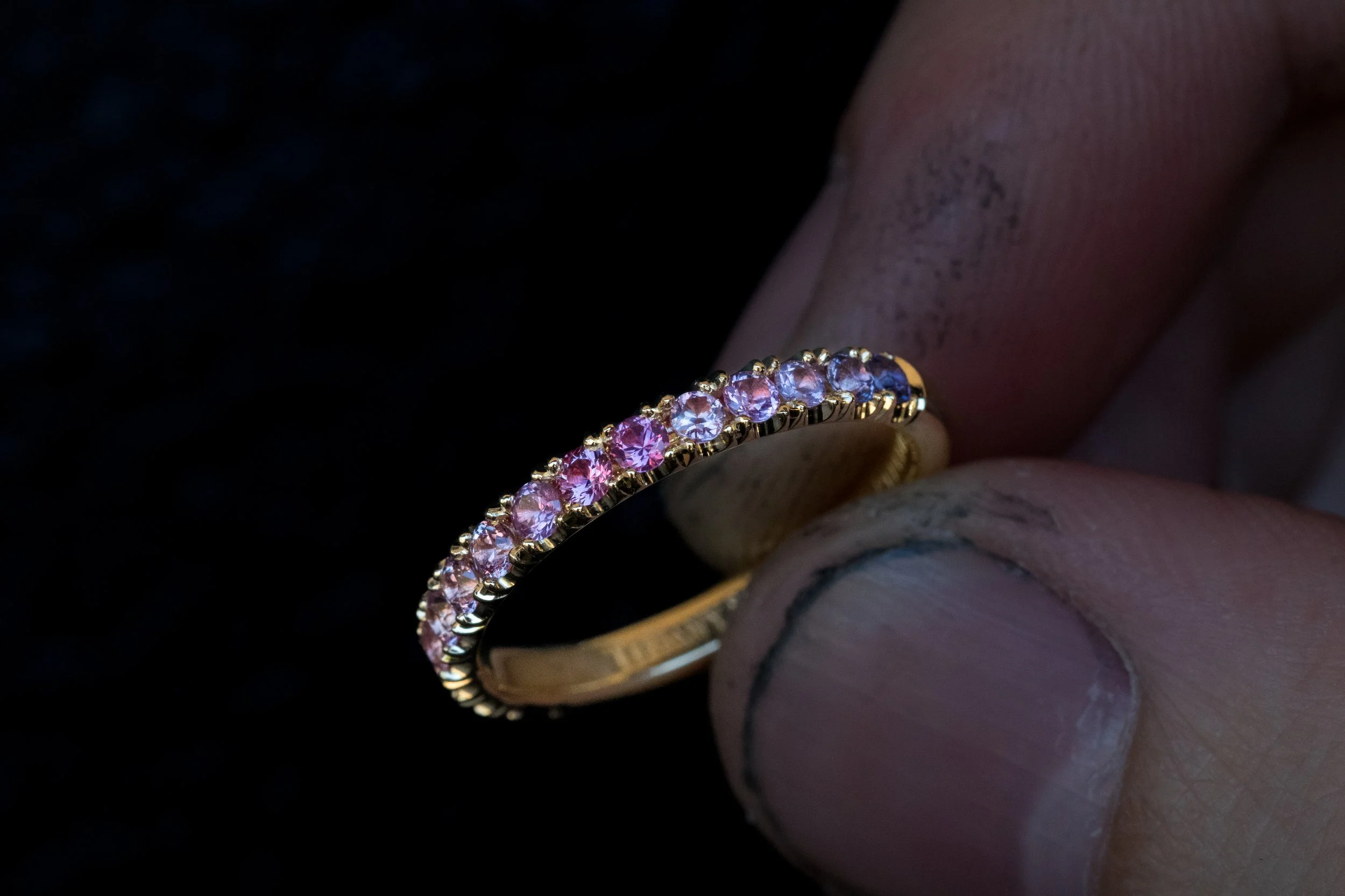 Close-up of a gold ring with pink and purple gemstones, held between fingers with visible dirt and nail imperfections against a dark background.
