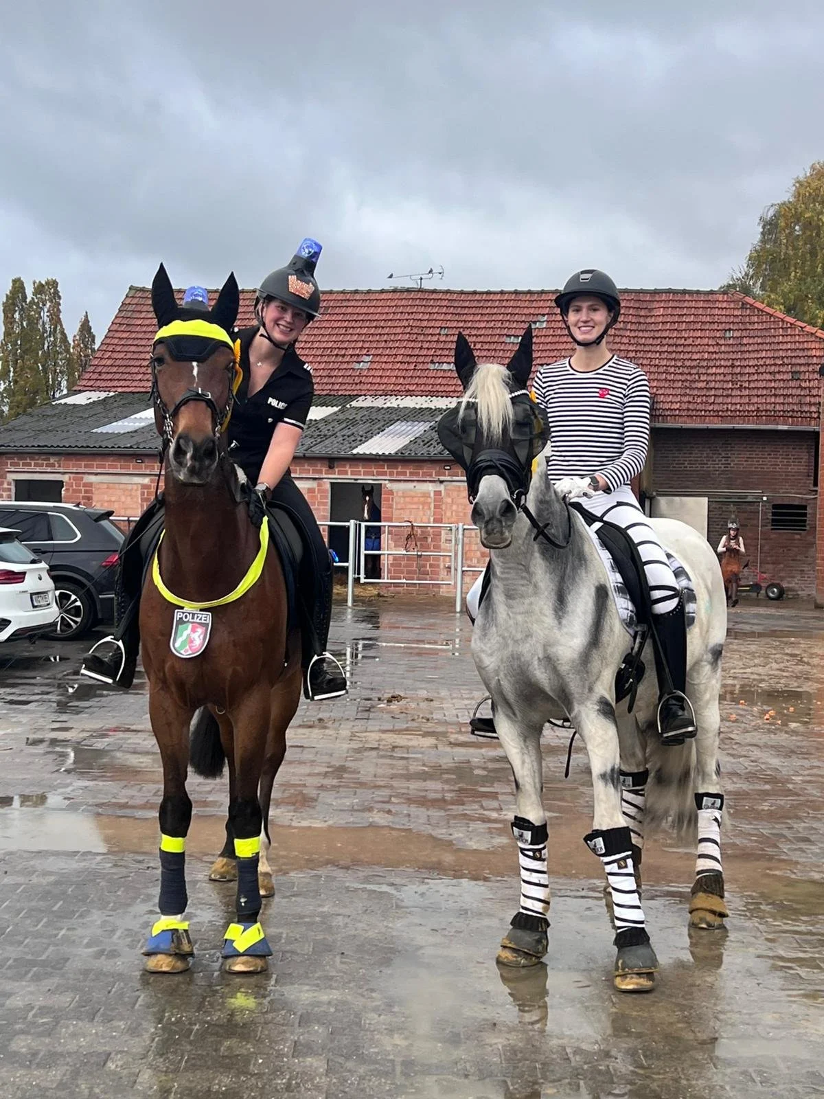 Zwei Frauen auf Ponys, eine in Polizeiuniform und die andere in Streifenmuster, im Regen, traurige Wolken, im Hintergrund ein Gebäude und Autos.