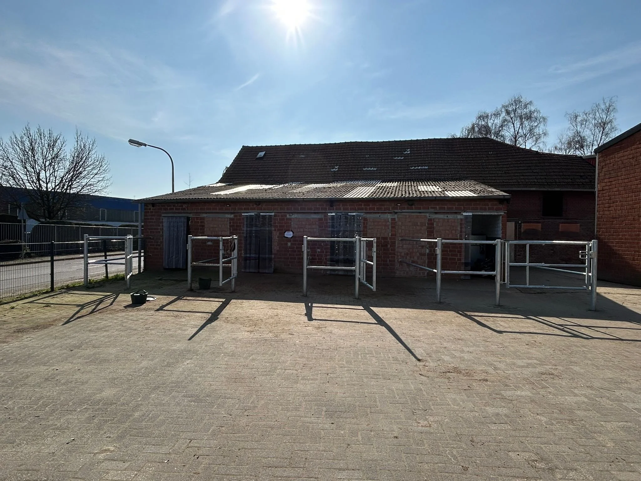 Ein kleines Gebäude mit rotem Backstein, davor eine abgezäunte offene Fläche mit Klettergerüsten, Schatten auf dem Boden, blauer Himmel, blattlose Bäume im Hintergrund.