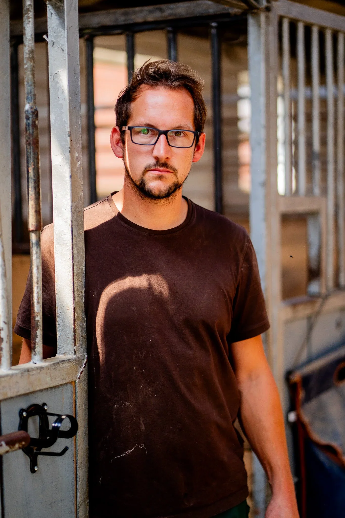 Junge Mann mit dunklem T-Shirt und Brille steht in einem Stall oder Gehege aus Metall, blickt ernst in die Kamera, im Hintergrund Holzwände und Metallgitter.