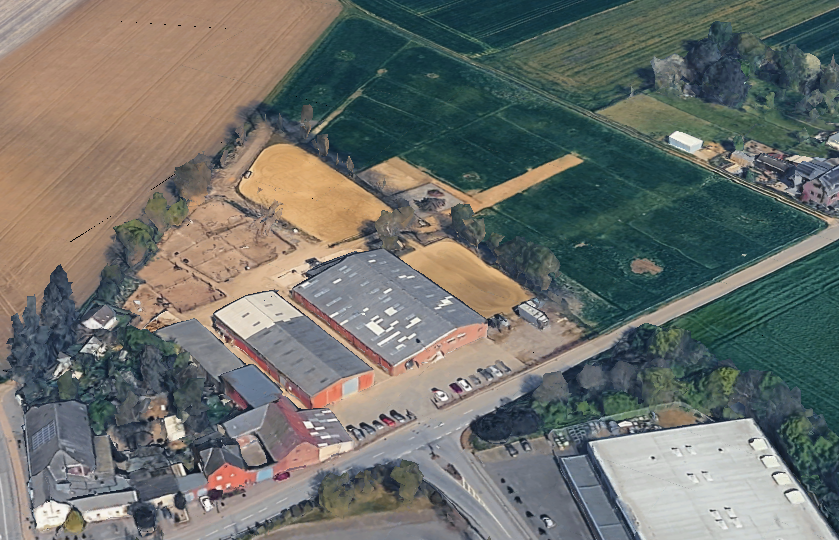 Luftaufnahme eines ländlichen Gebiets mit landwirtschaftlichen Feldern, einem Hof mit mehreren Gebäuden und Parkplätzen, umgeben von grünen Feldern und Bäumen.