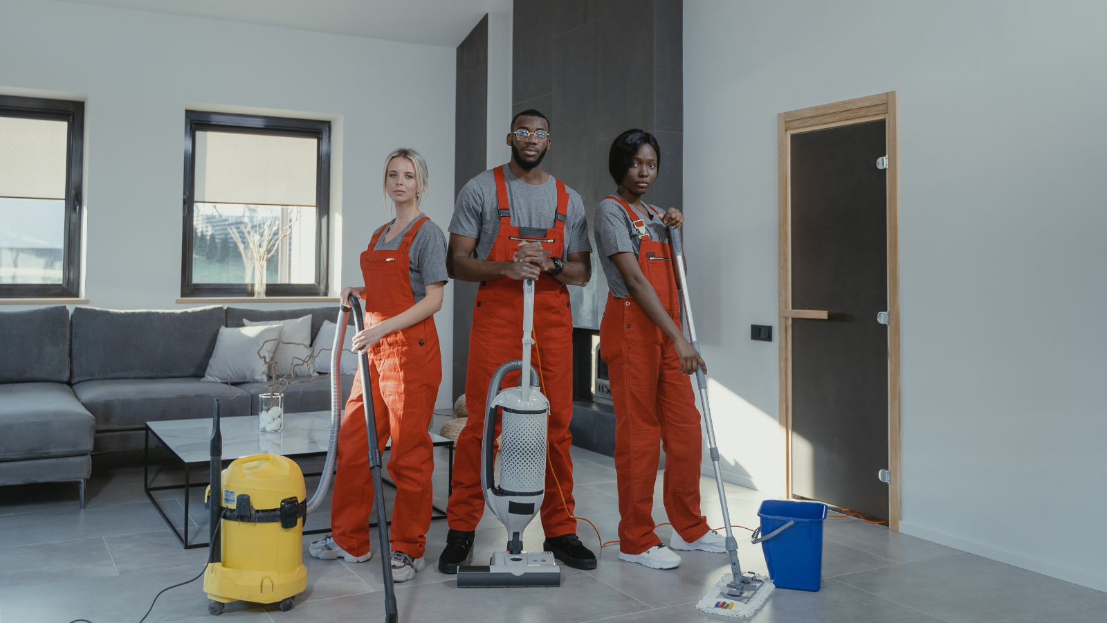 Three professional cleaners in matching uniforms vacuuming and mopping an indoor living room with modern decor.