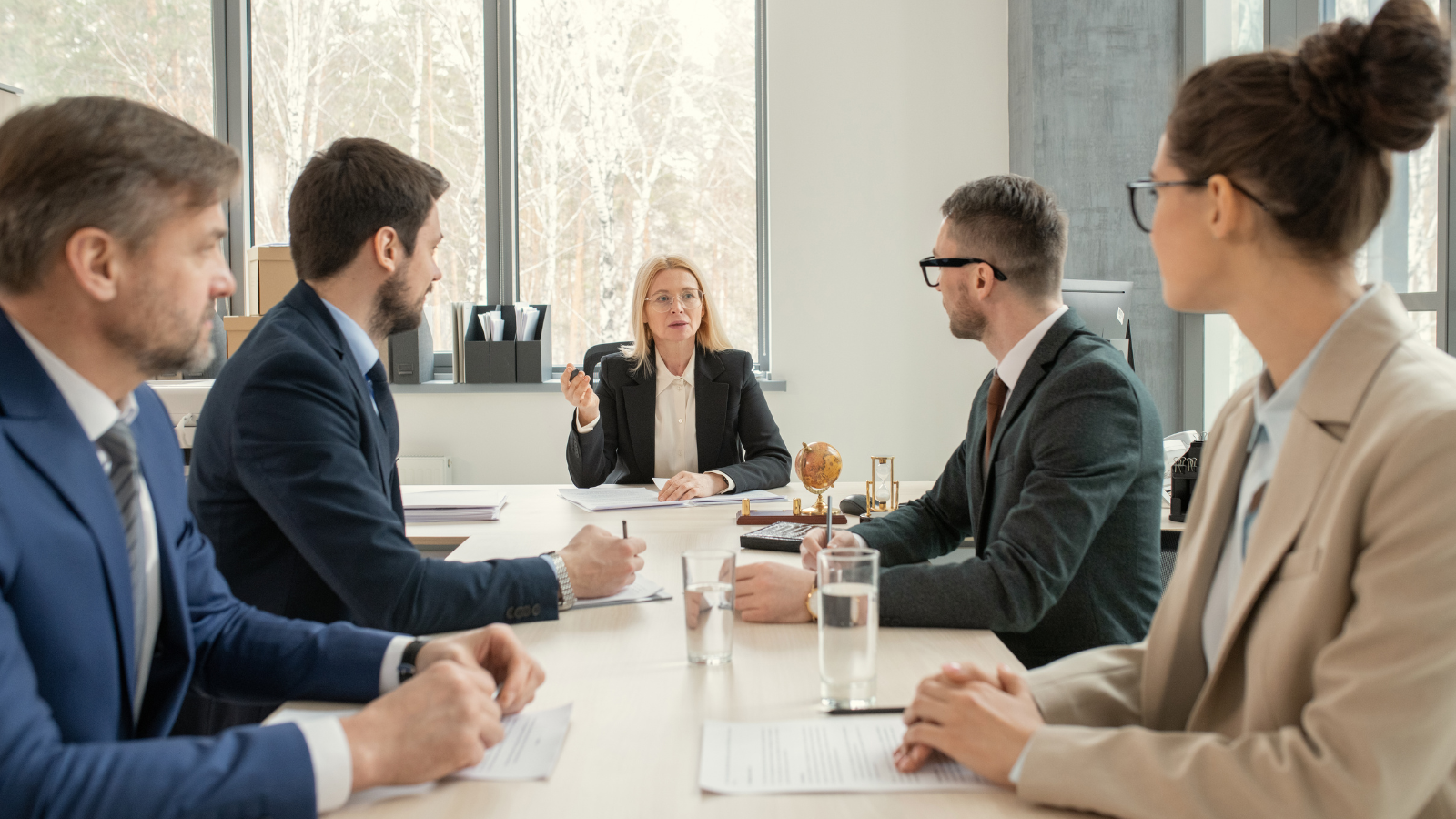 Business meeting with five professionals in a modern conference room with large windows, engaging in discussion around a table.