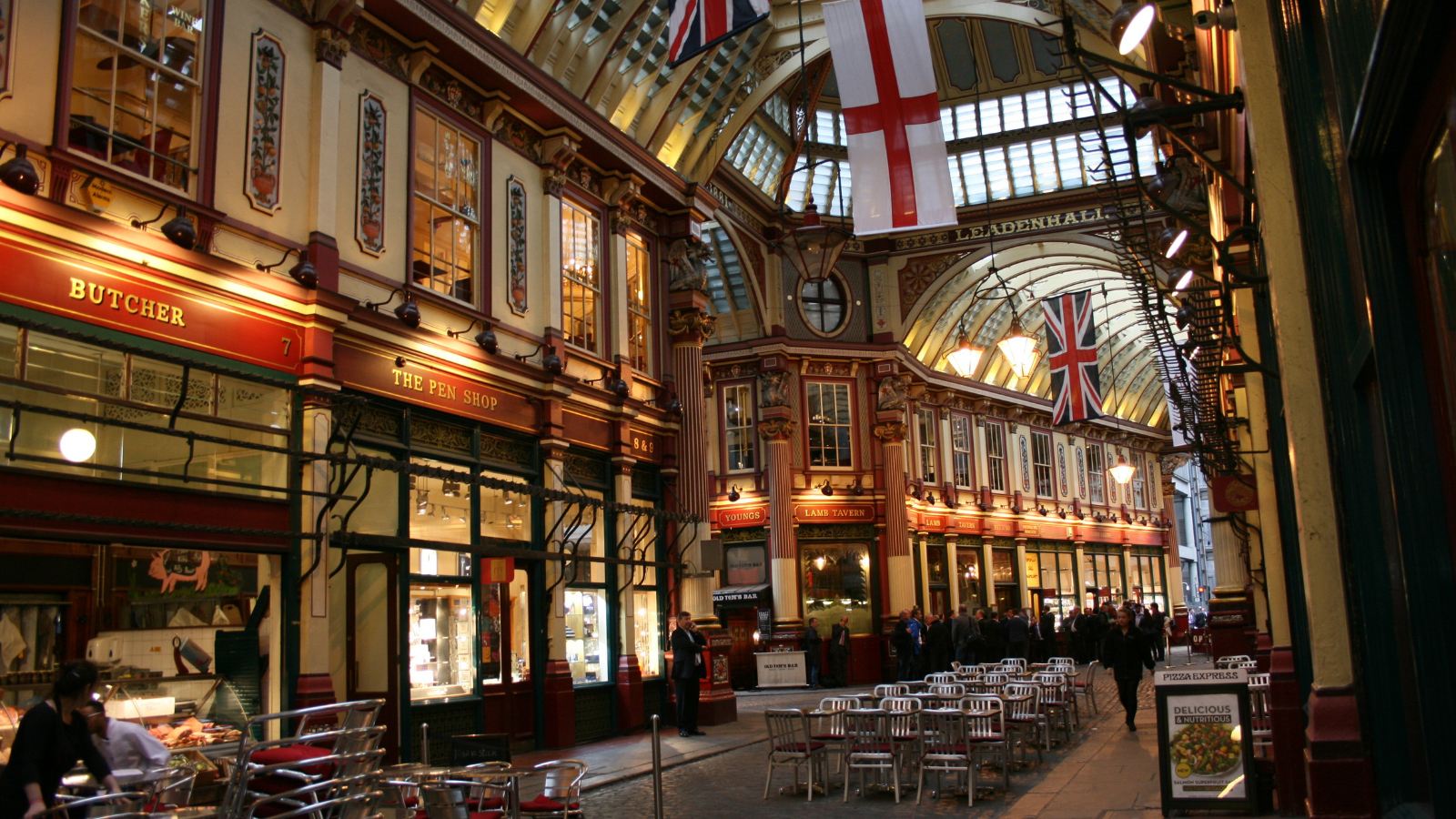 Interior of a historic shopping arcade with ornate architecture, flags, and several shops and cafes, including Butcher and The Pen Shop, with people walking and sitting outside.