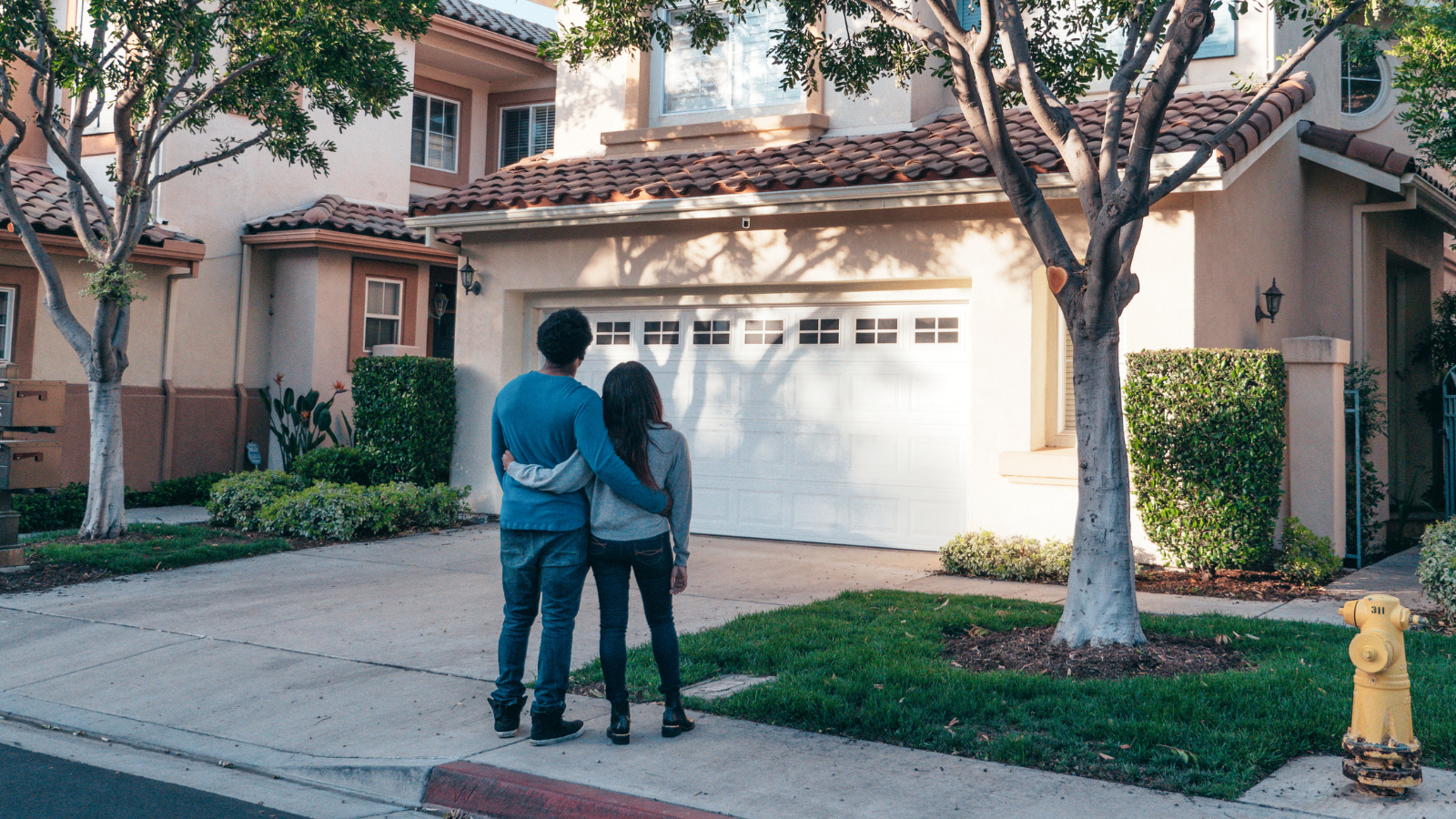 A young couple stands outside a home in a suburban neighborhood, viewing the house. The house features a white garage door, stucco walls, and a tiled roof, with trees and bushes surrounding it.