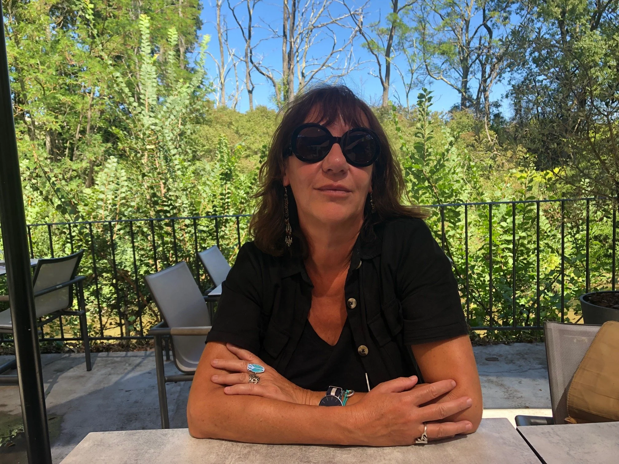 Une femme assise à une table en extérieur, portant de grosses lunettes noires, une tenue noire, avec des bijoux, dans un environnement verdoyant avec un ciel bleu en arrière-plan.
