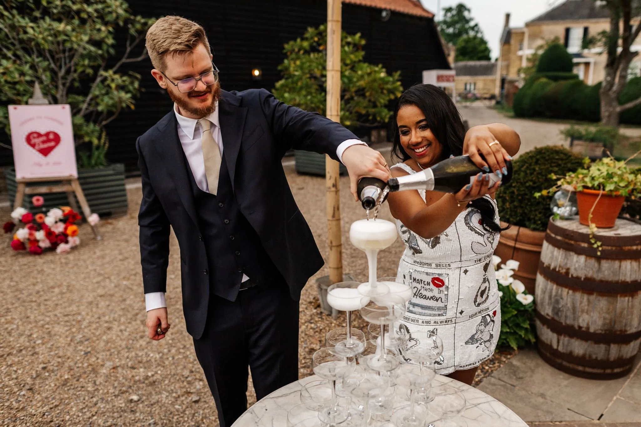 Diana and Sean pouring a champagne tower with their pink heart wedding welcome sign behind them