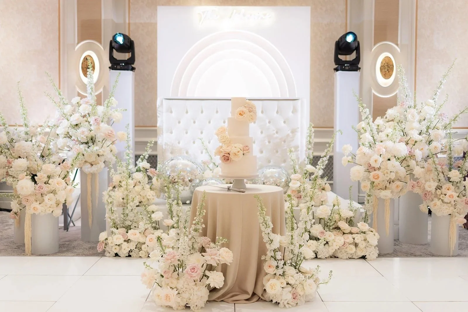 Elegant wedding cake with flower decorations at the center, surrounded by white and blush pink floral arrangements in tall vases on white pedestals, set against a white tufted backdrop with soft lighting.