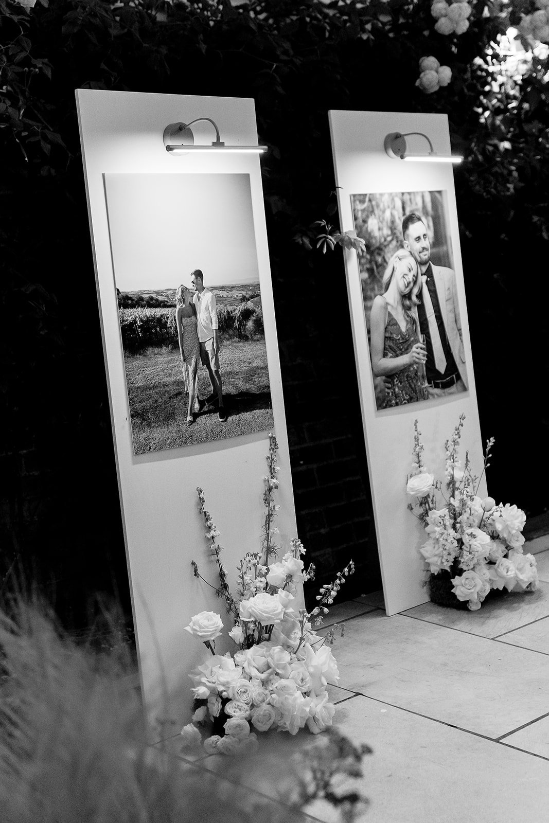 Two photo display boards decorated with flowers, each illuminated by a small light, showing black-and-white photos of couples at a wedding or special event.