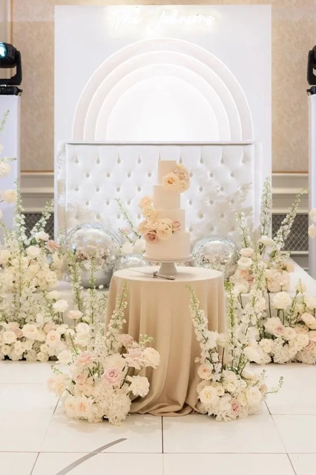 Elegant wedding cake with flowers on a beige table, surrounded by white and blush floral arrangements, set against a soft white backdrop with a tufted white panel.
