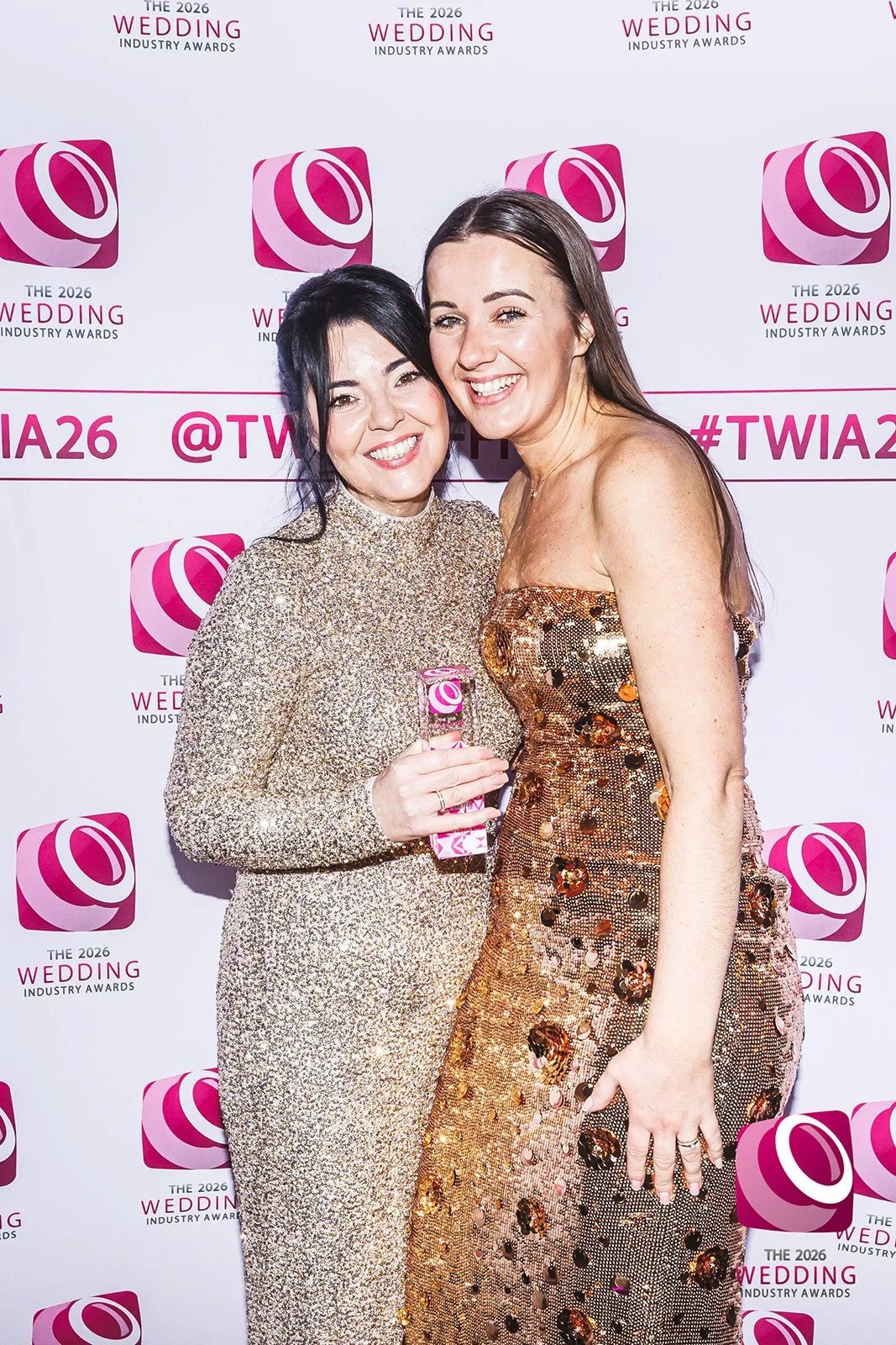 Two women in elegant dresses posing together at the 2026 Wedding Industry Awards, smiling for the camera with a pink and white backdrop featuring the event's logo.