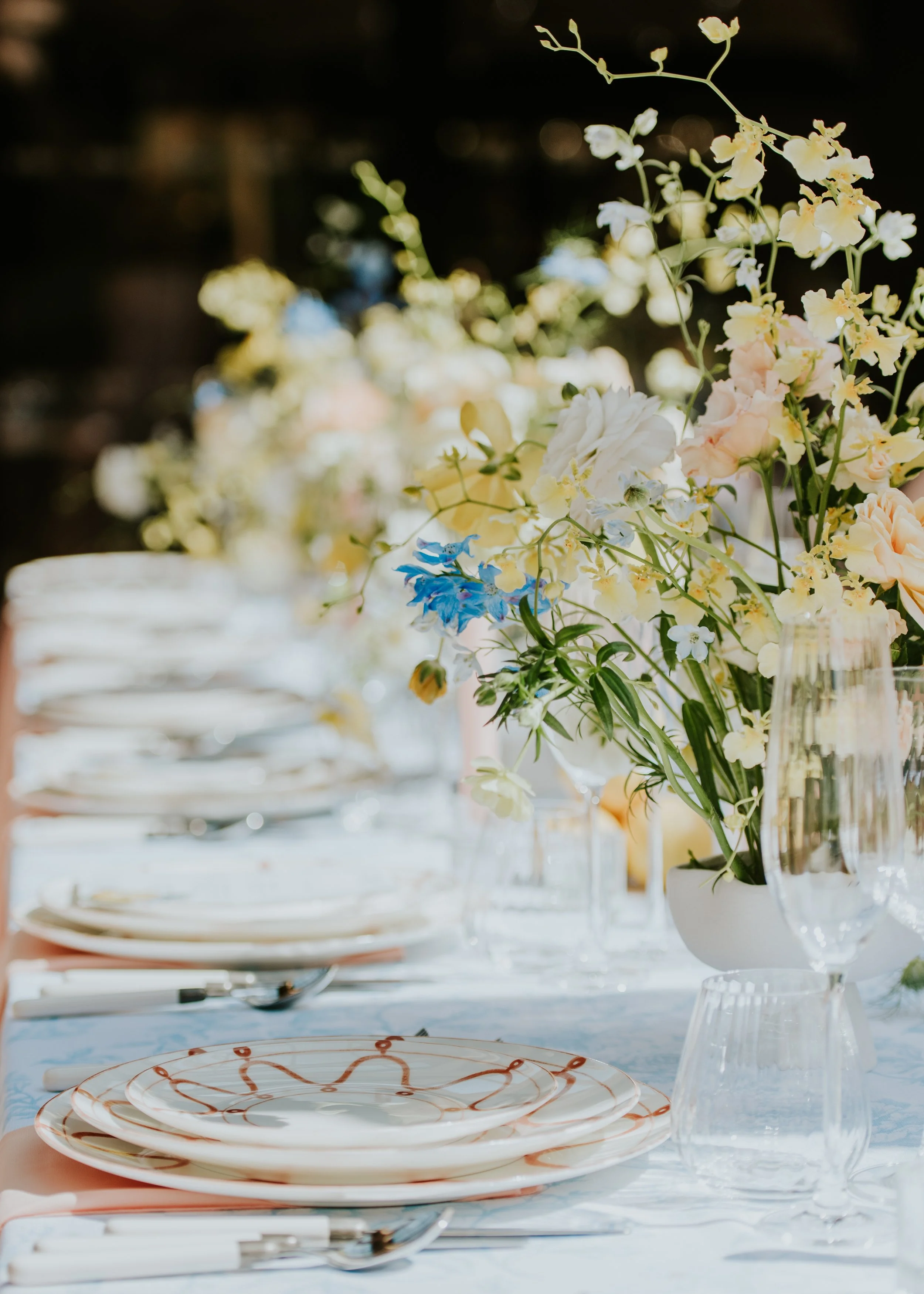 Elegant dining table set with floral arrangements, white plates with orange and brown patterns, wine glasses, and silverware in a softly lit environment.
