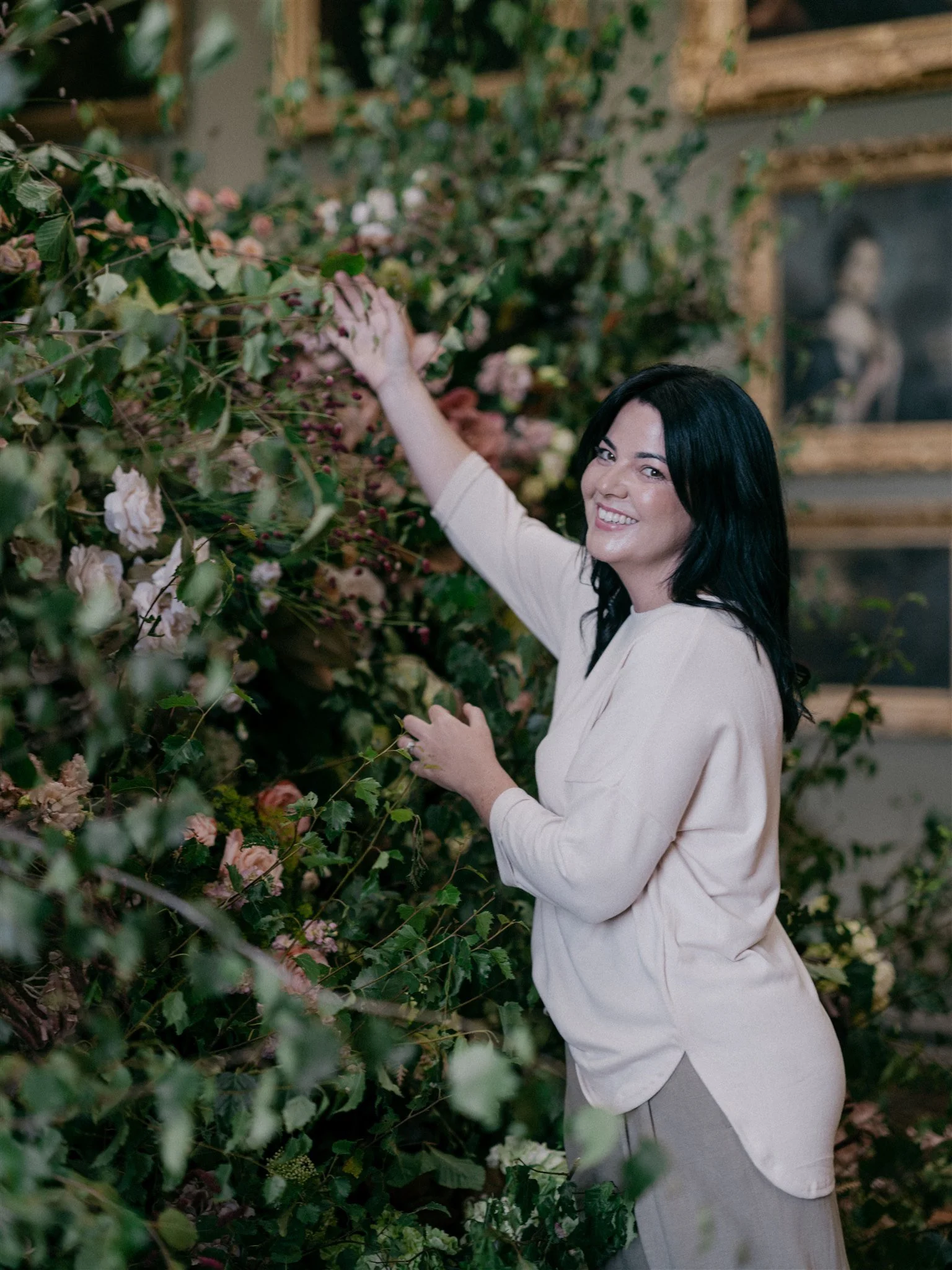 Woman with black hair smiling and wearing a light-colored top, touching pink and white flowers on a lush green shrub in a garden or greenhouse setting.
