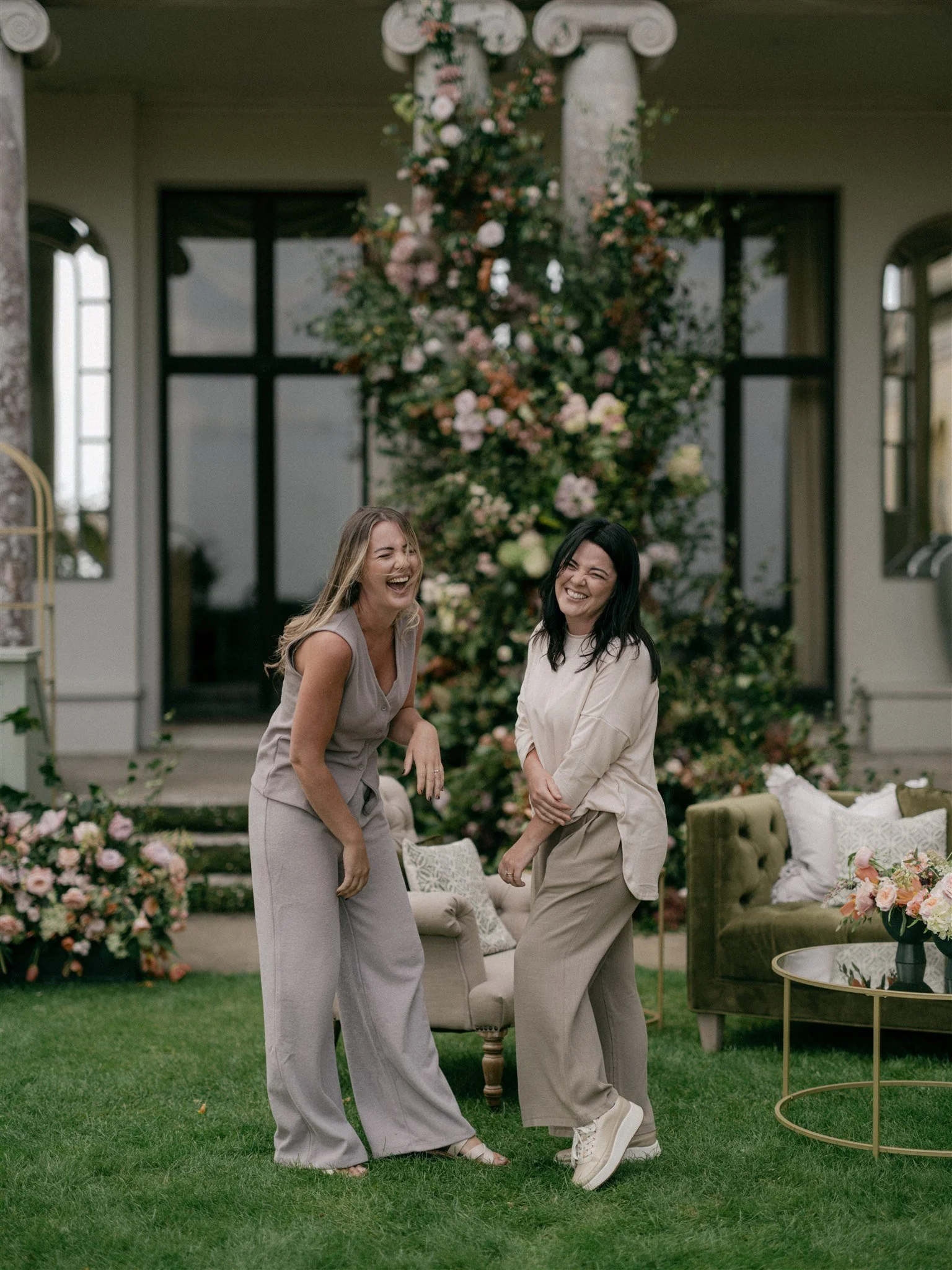 Two women laughing and standing on a green lawn in front of a floral decorated background with chairs, a sofa, and a table with a flower arrangement, at an outdoor event.