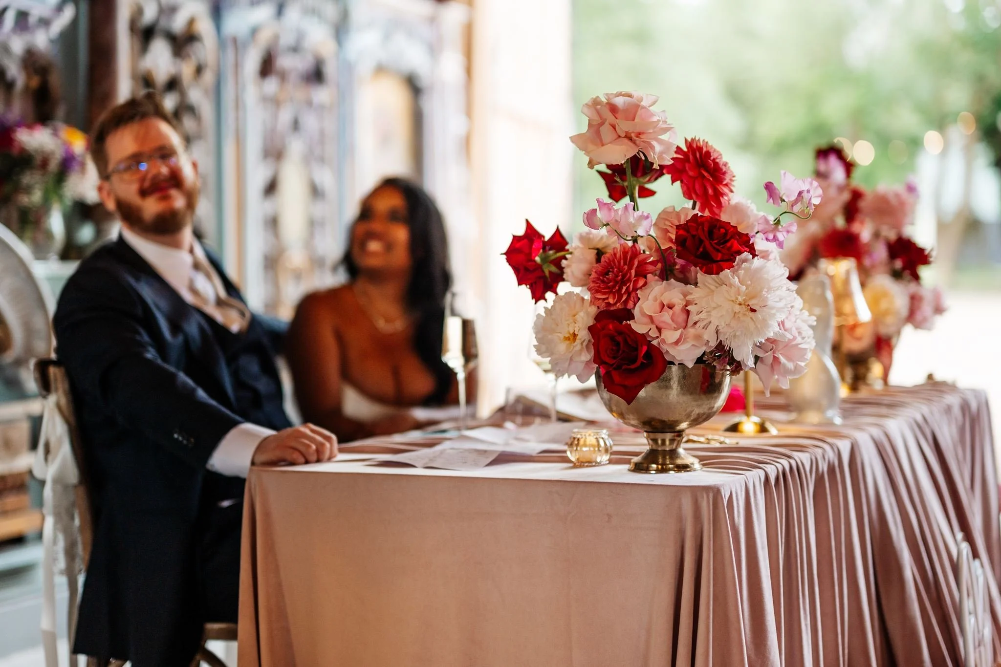 Diana and Sean seated behind pink and red wedding flowers at their blush-draped reception table