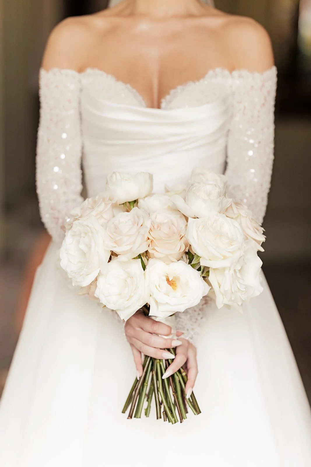 A bride holding a bouquet of white roses and peonies, wearing a white wedding dress with lace sleeves.