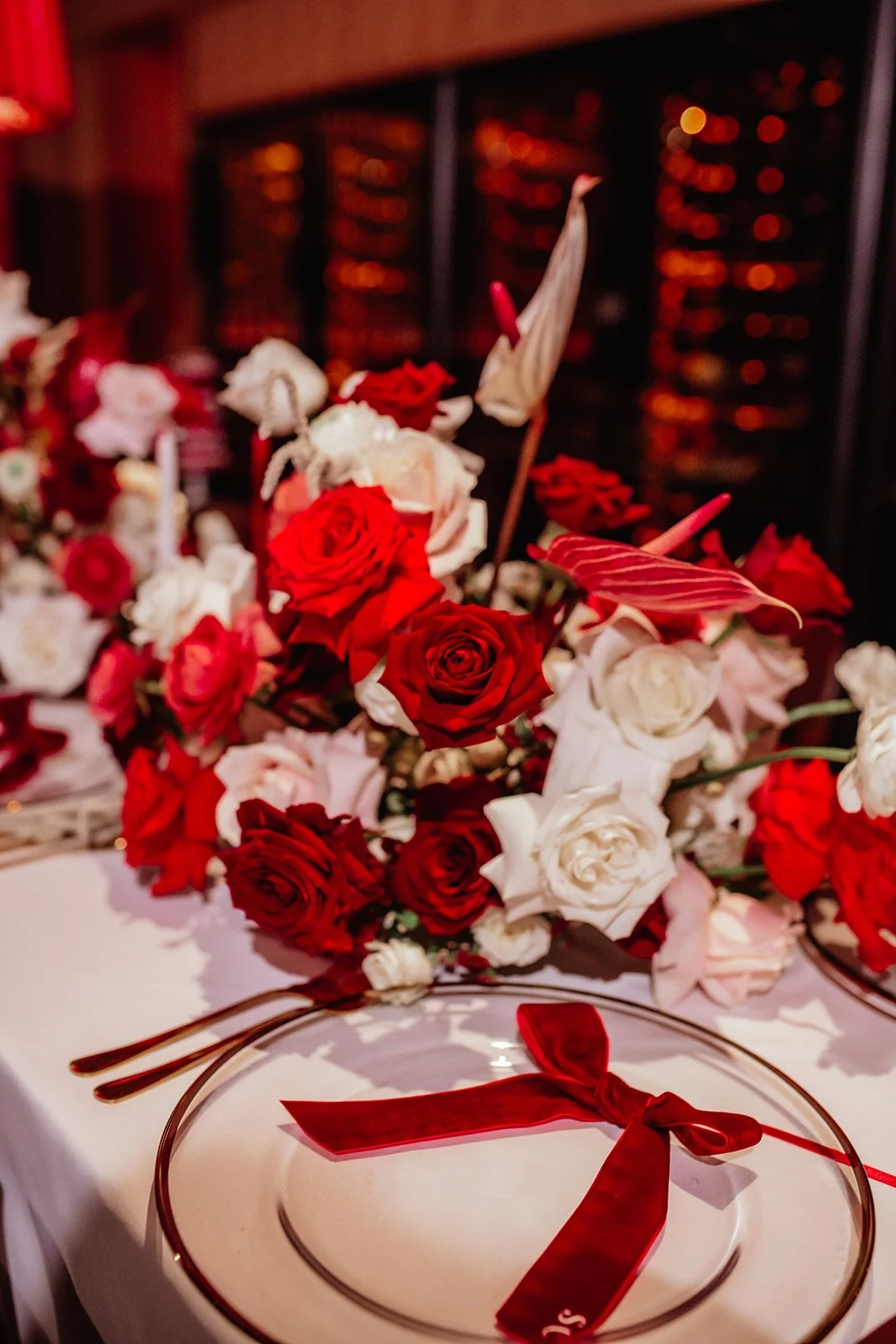 A table centerpiece made of red and white roses with an abstract modern arrangement, with a clear glass plate with a red ribbon in front, set in a warmly lit room.