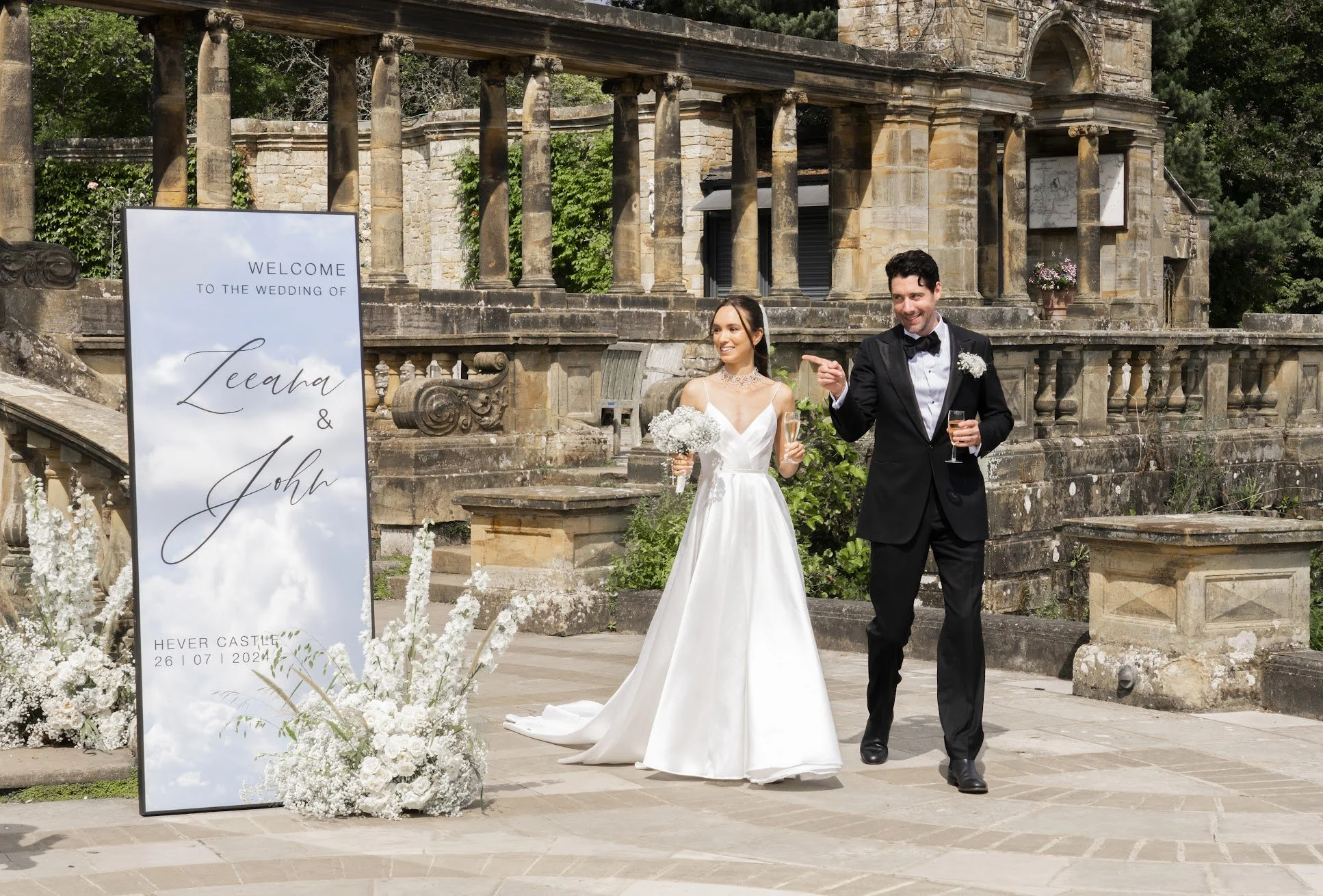 Couple beside a mirrored wedding welcome sign and white floral displays at Hever Castle