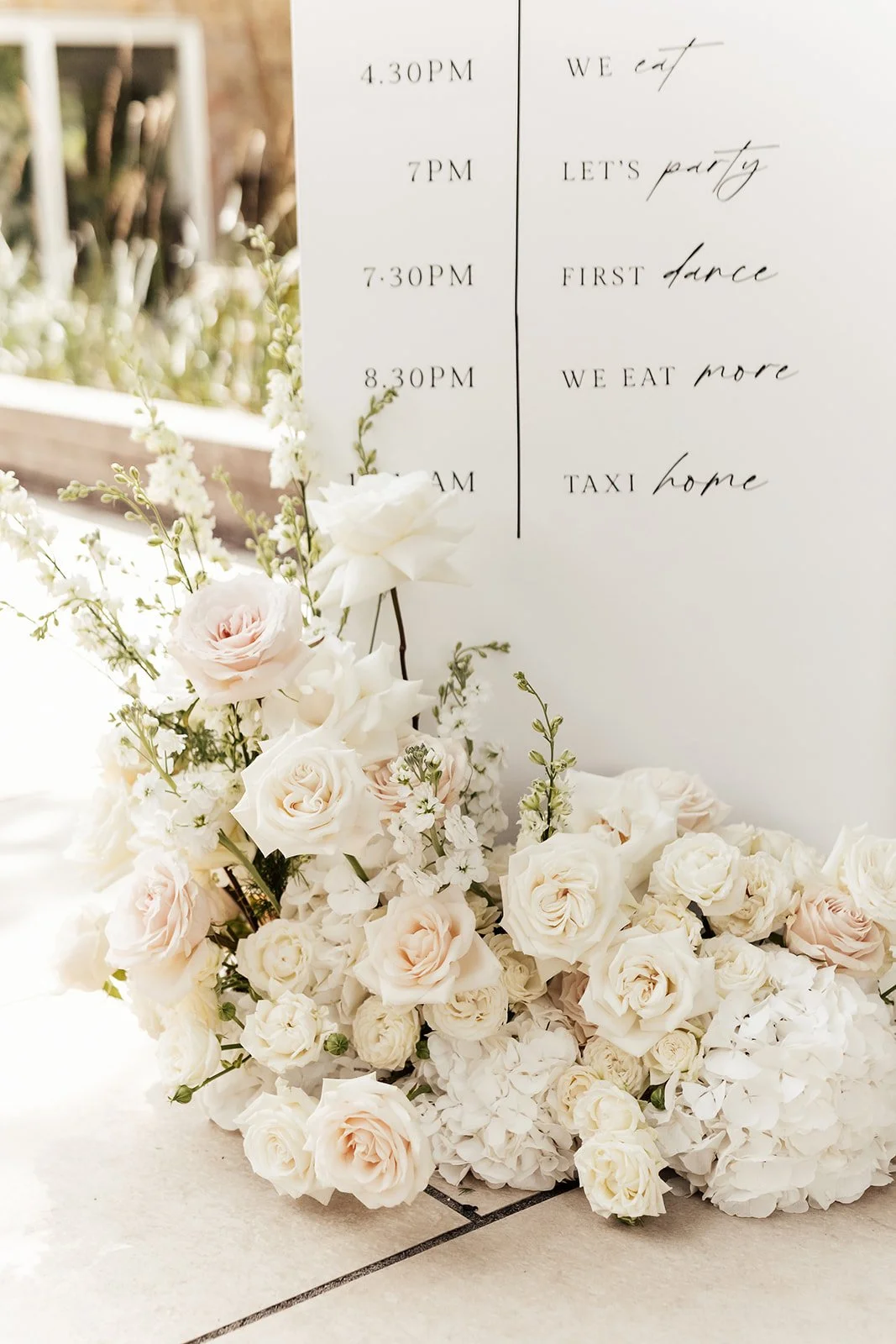 A schedule on a whiteboard with times and events, accompanied by a floral arrangement of white and pale pink roses and other white flowers at the base.