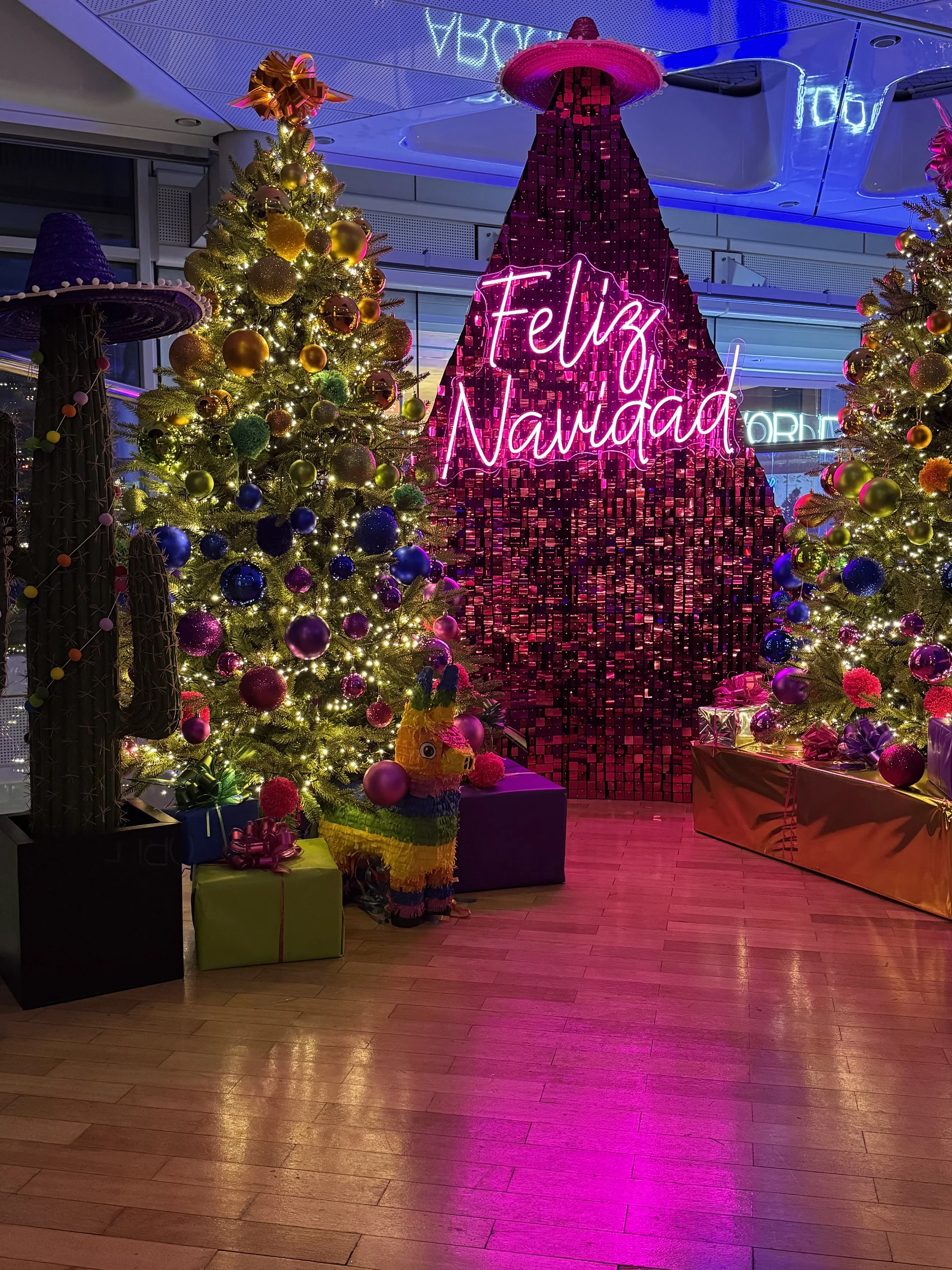 Colorful Christmas display with decorated trees, a piñata, wrapped presents, and a neon sign that reads 'Feliz Navidad' in pink, with a pink and purple background.