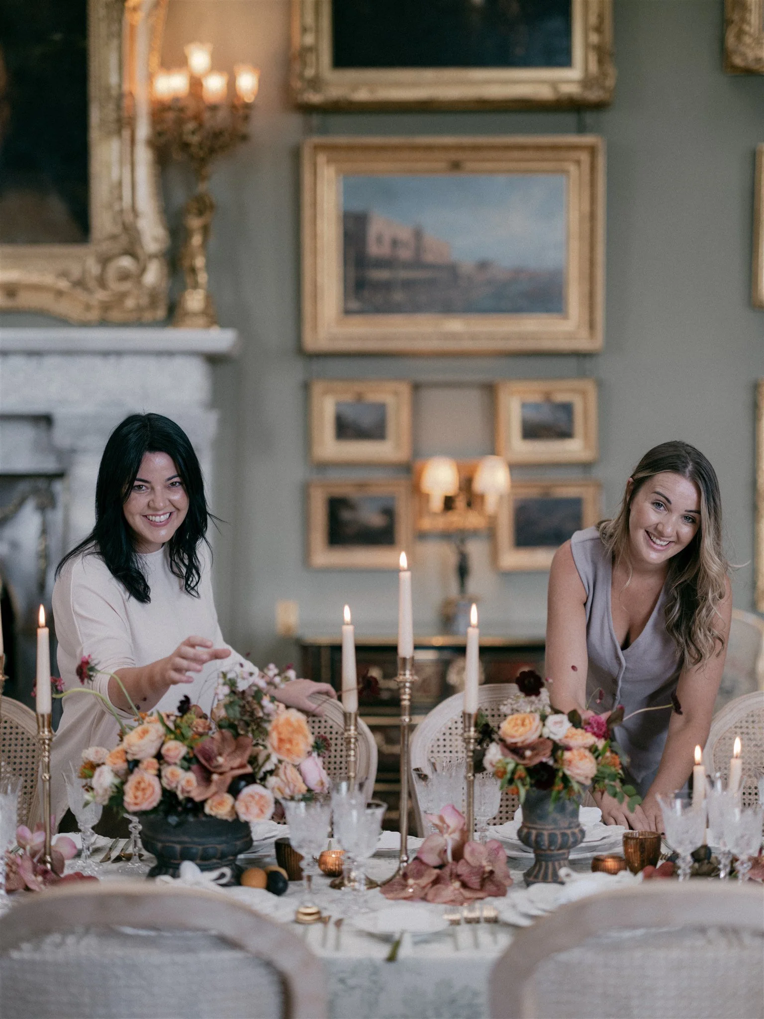 Two women smiling and setting a decorated dining table with candles and floral centerpieces in an elegant room with framed paintings and a fireplace.