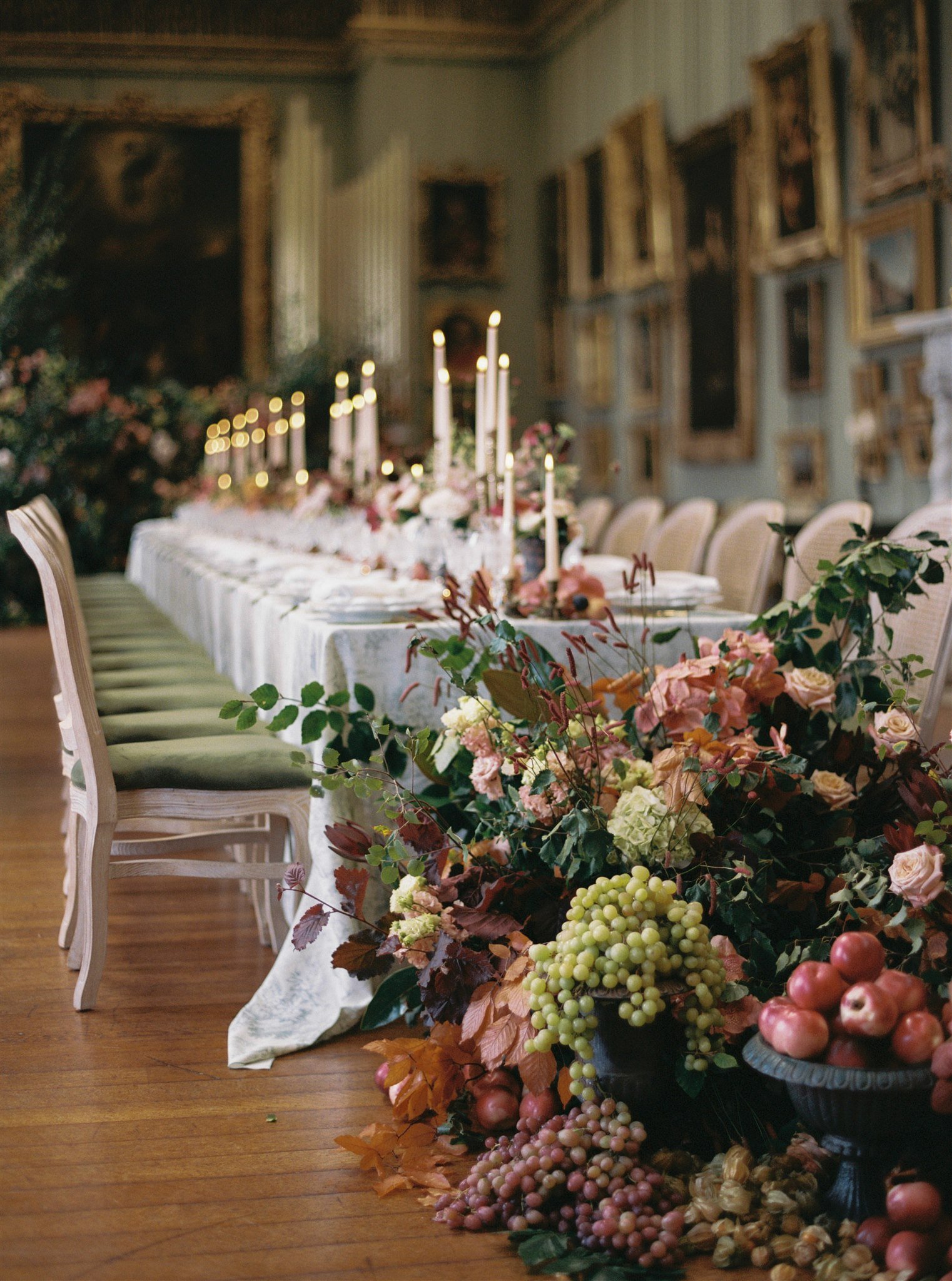 A long dining table decorated with candles, floral arrangements, and fruit, set in an elegant room with framed paintings and ornate moldings.