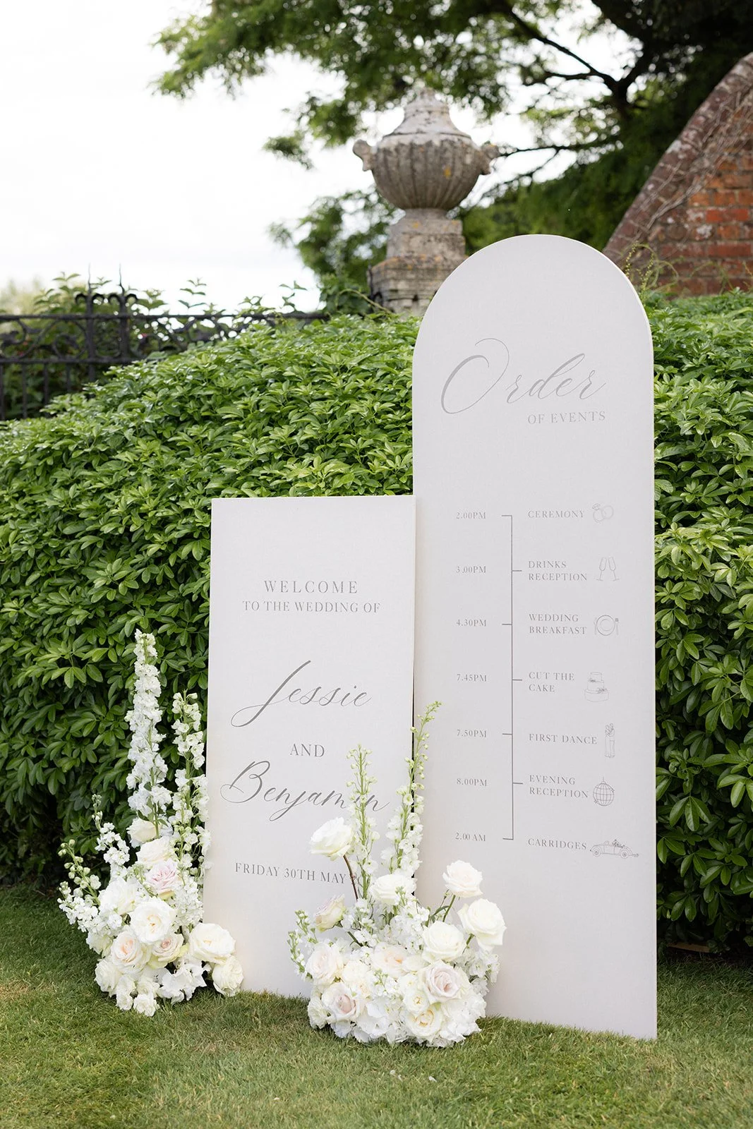 Wedding signage with schedule and welcome message surrounded by white flowers, set outdoors with greenery and decorative stone urns in the background