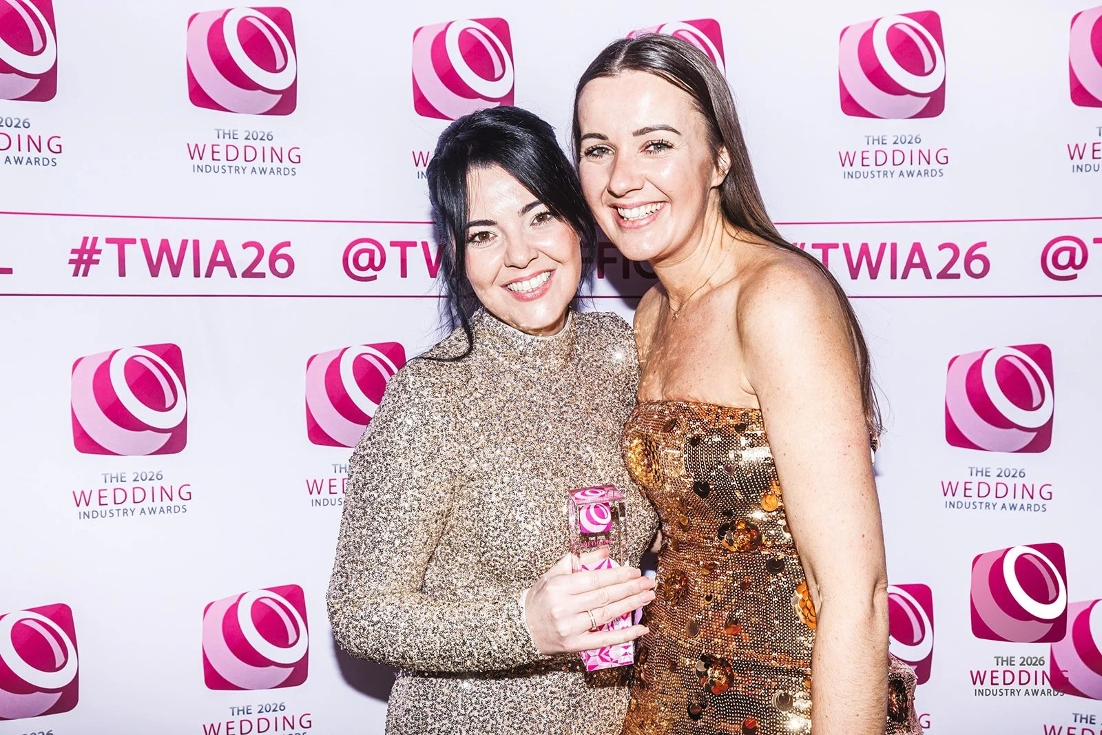 Two women smiling at an awards event, one holding a pink trophy, standing in front of a backdrop with logos and the hashtags #TWIA26.