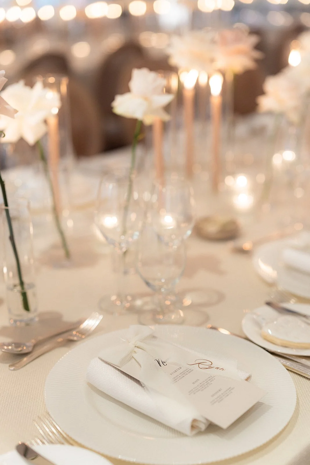 Elegant table setting with white napkin and menu on a dinner plate, surrounded by wine glasses and cutlery, decorated with white flowers and candlelight.